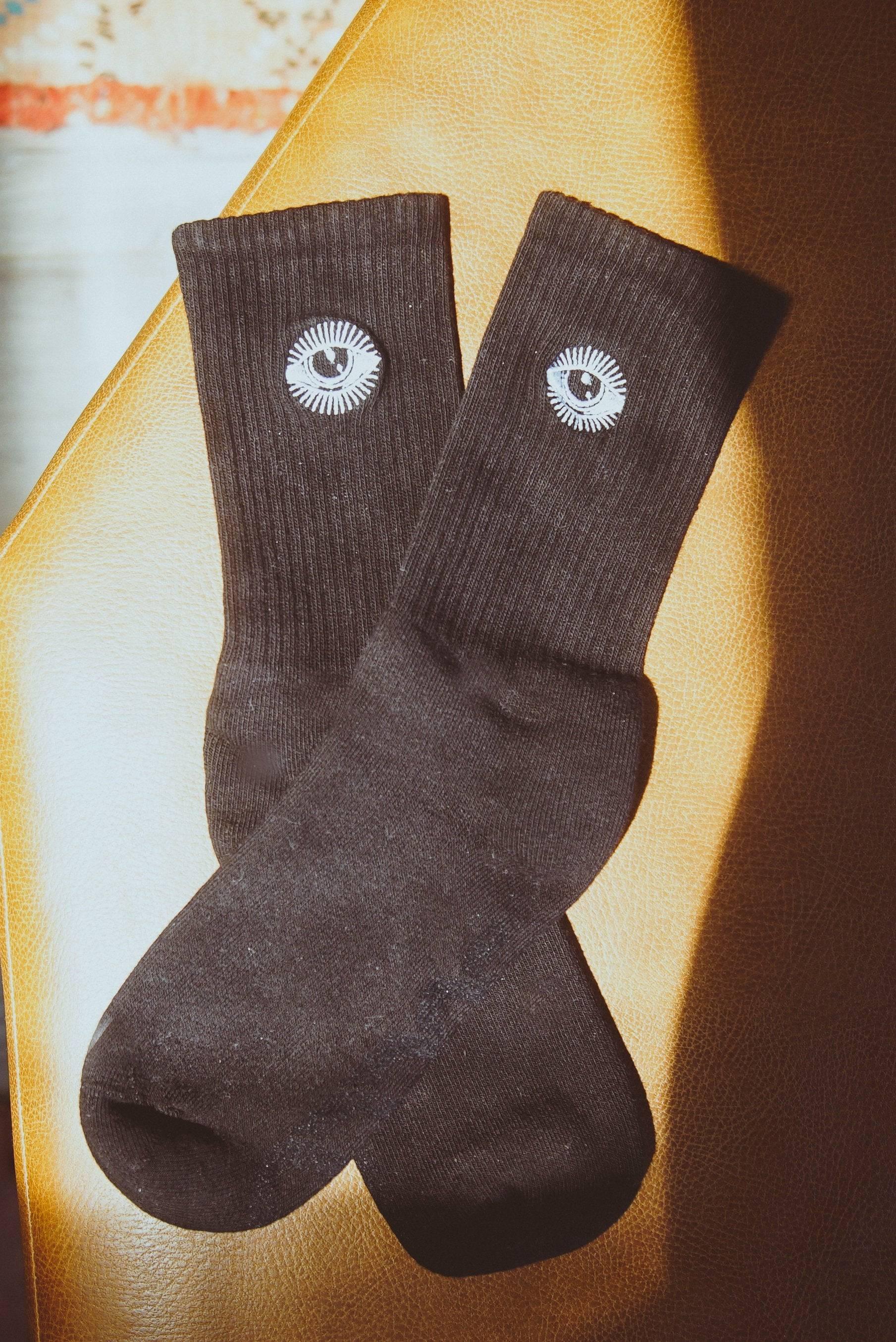 Black crew socks with white eye embroidery on ribbed cuff, placed on brown leather surface