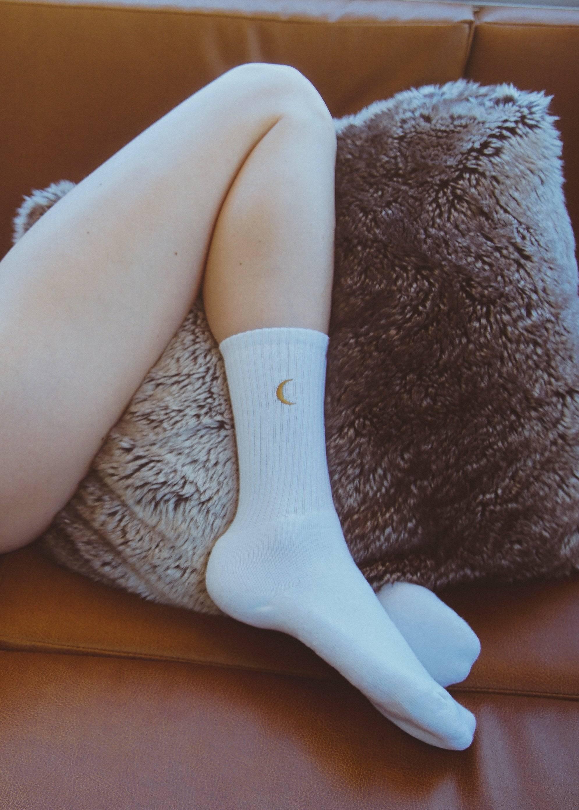 Person wearing white ribbed socks with a small crescent moon, lying on brown leather couch with furry pillow