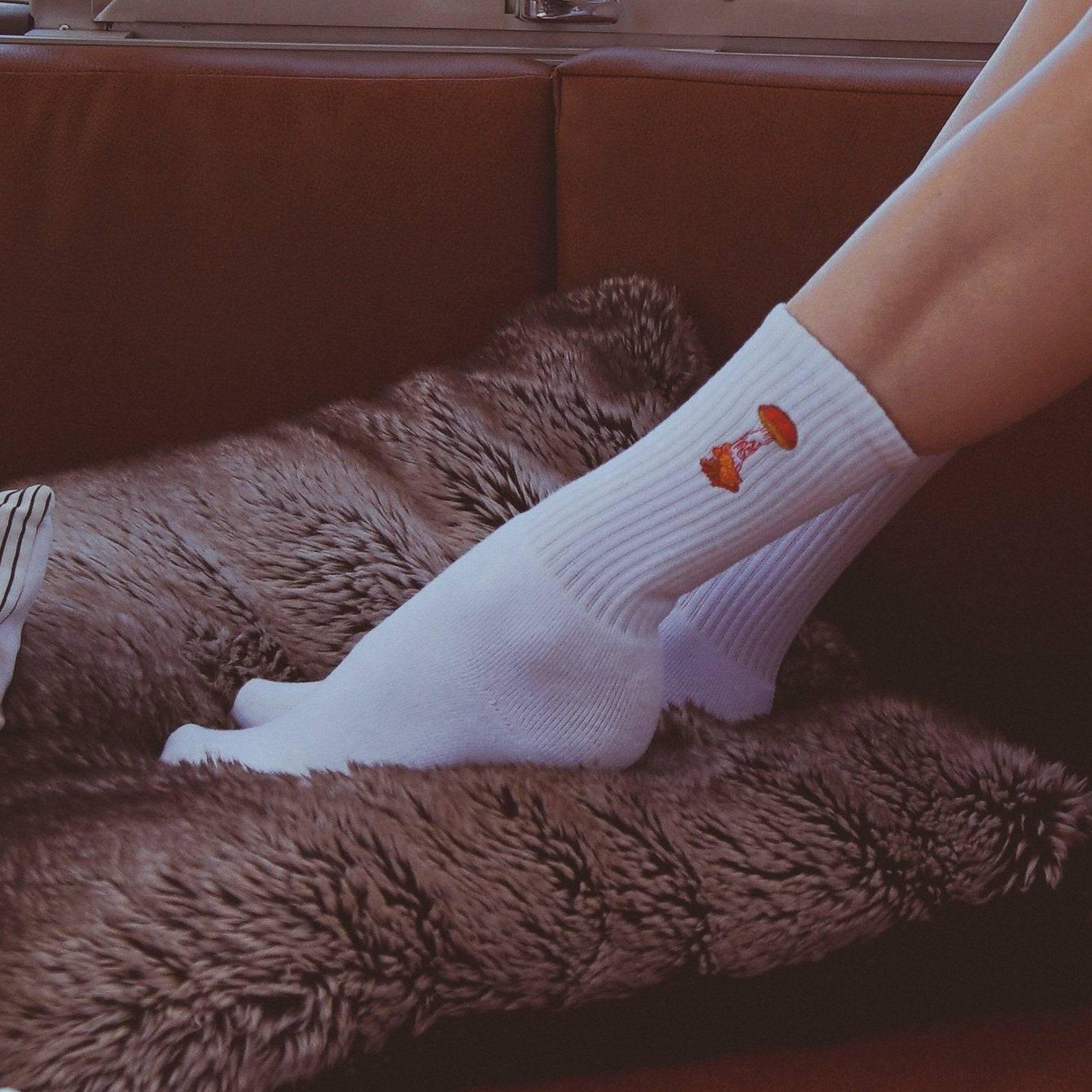 White ribbed socks with a small orange mushroom design worn on feet resting on a soft brown furry blanket on a brown couch