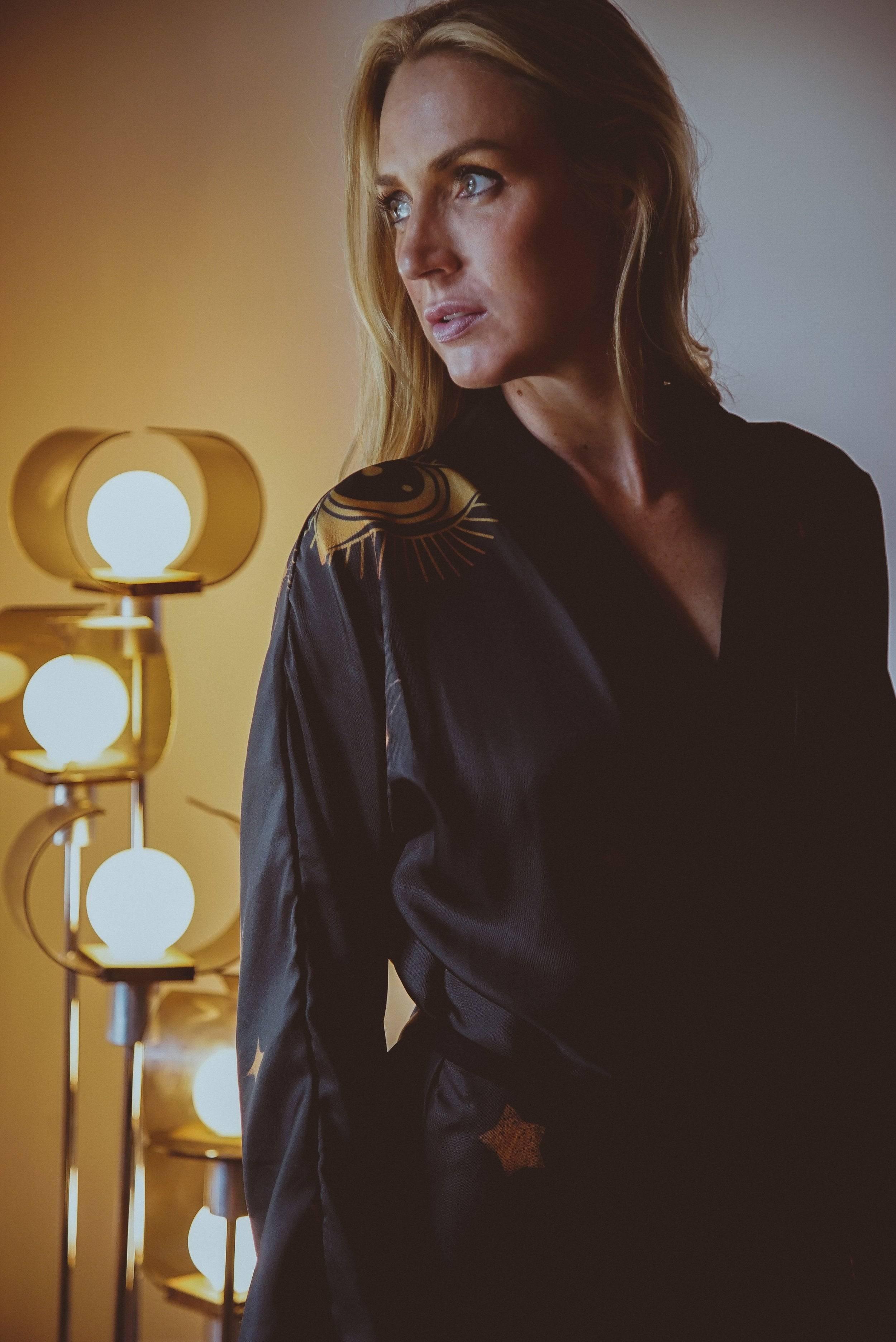 Blonde woman in black robe with sun and star patterns, standing near glowing modern floor lamp