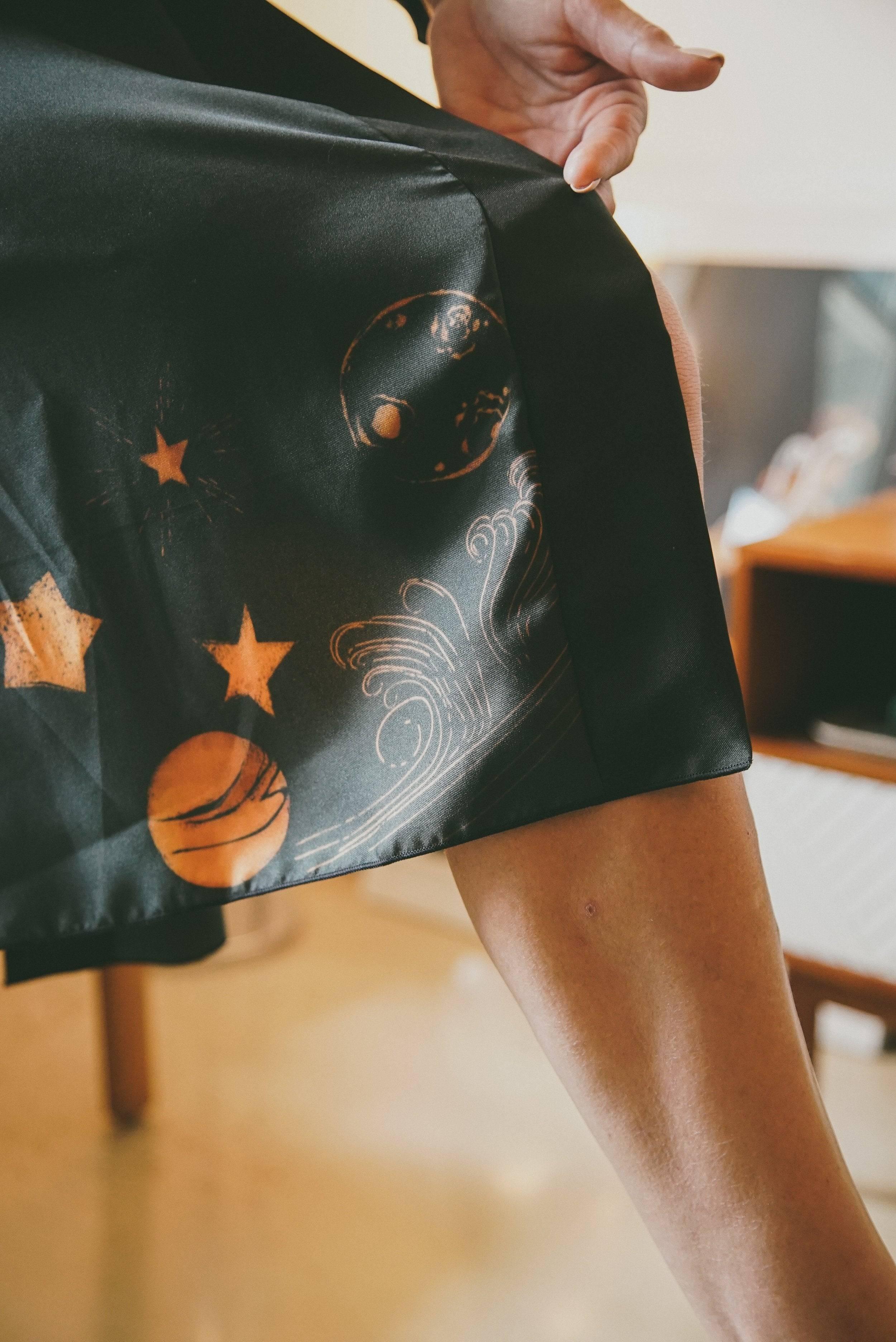 Hand lifting black fabric with orange celestial and wave designs, showing a person's leg indoors
