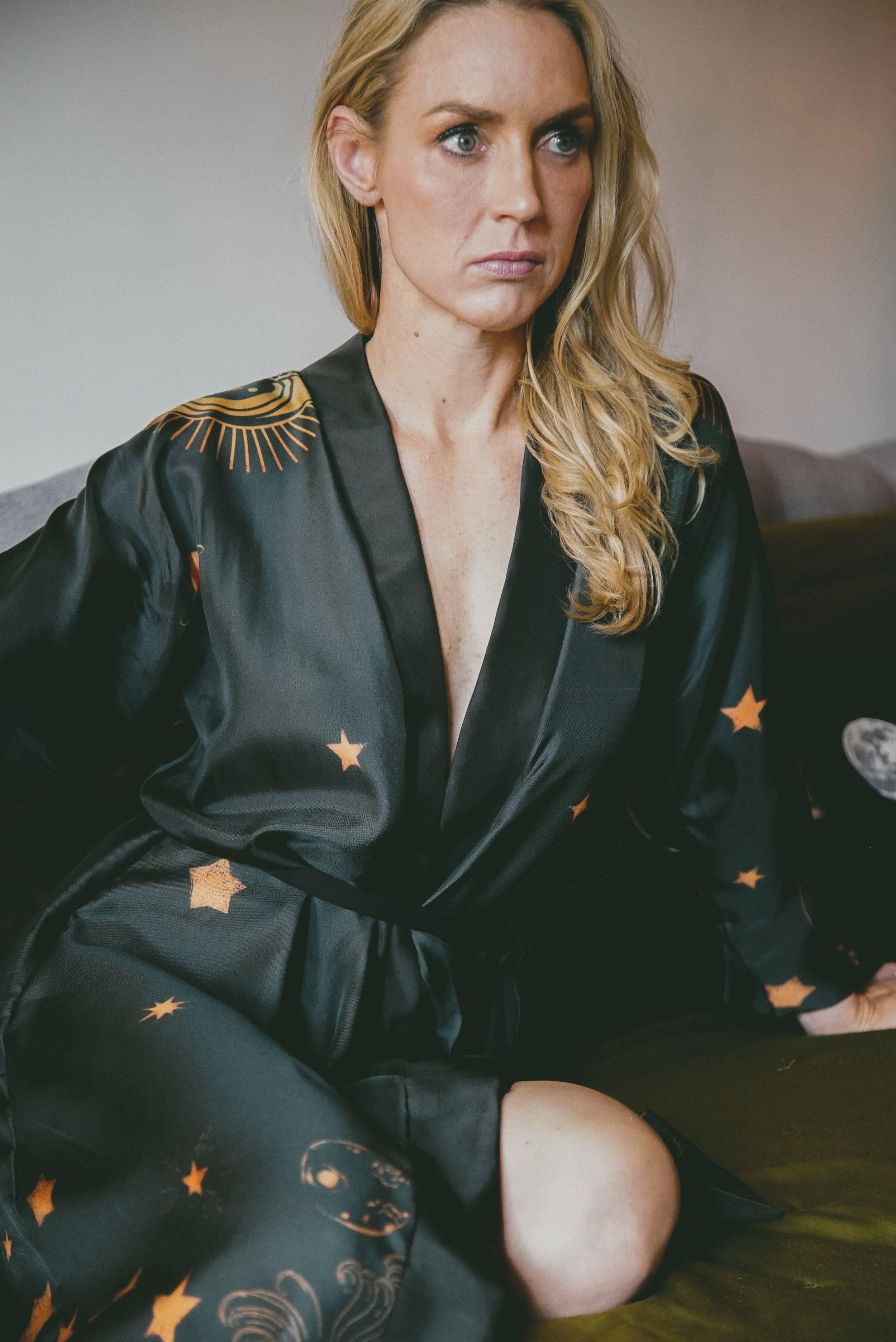 Blonde woman wearing black satin robe with gold celestial sun and star patterns sitting on green couch
