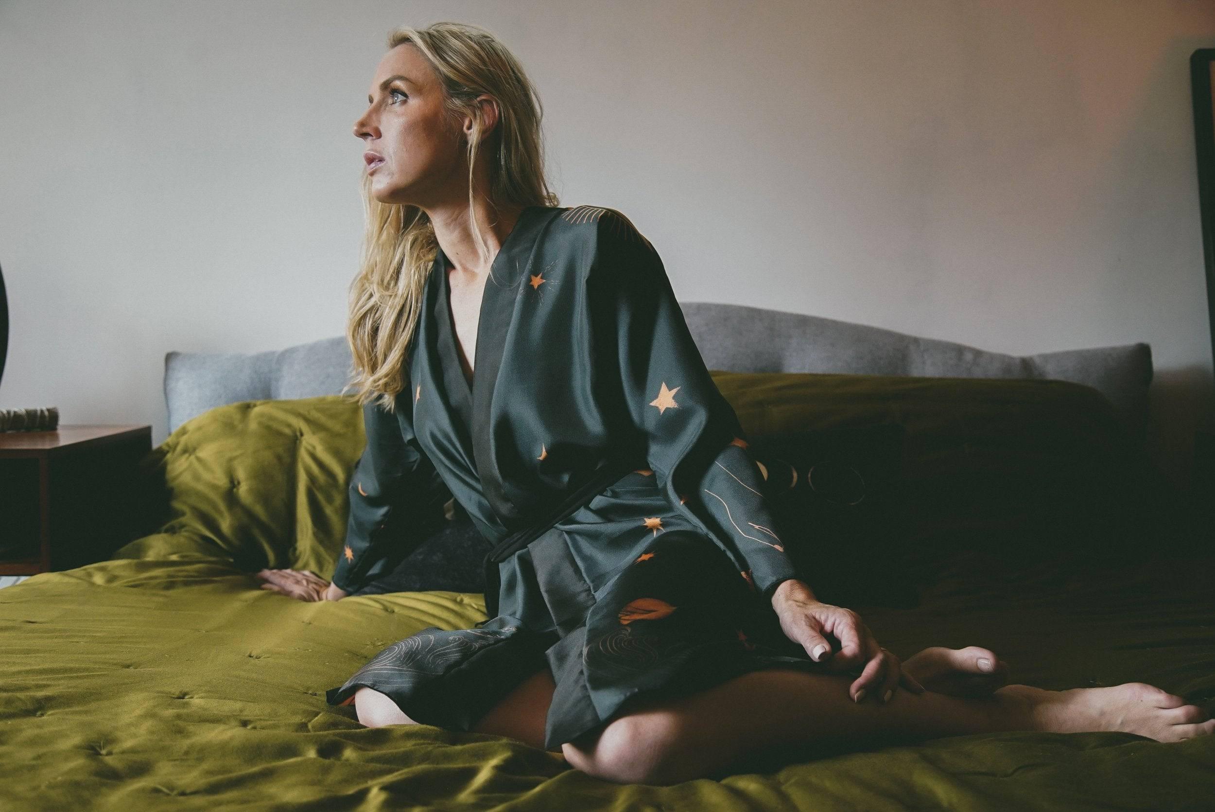 Blonde woman in dark star-patterned robe sitting on olive green bed in softly lit bedroom