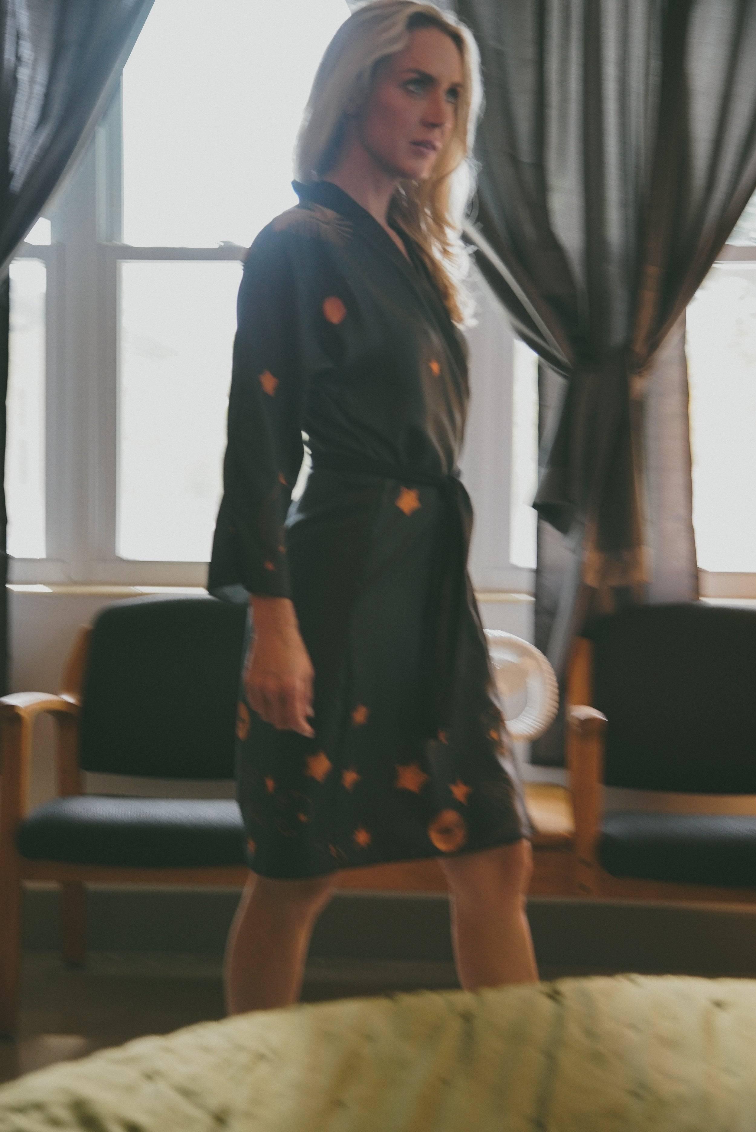 Blonde woman in black robe with orange star and planet patterns walking indoors near chairs and windows