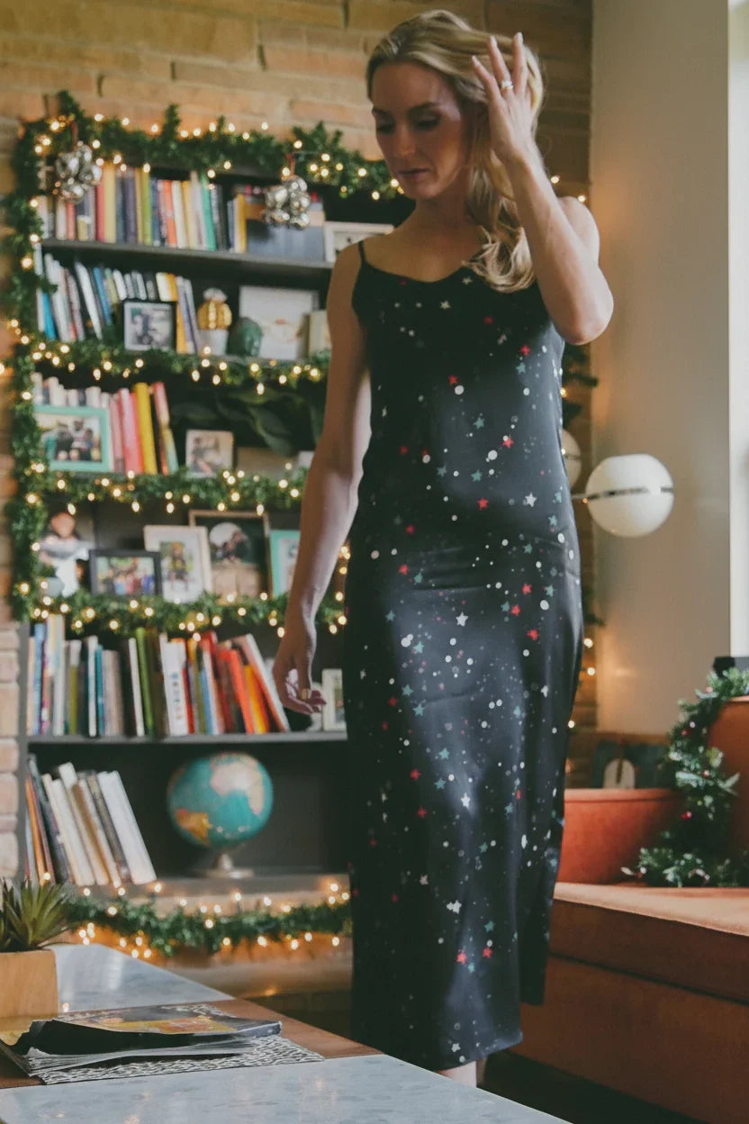 Woman in black star-patterned dress standing in a cozy, decorated living room with bookshelves and holiday lights