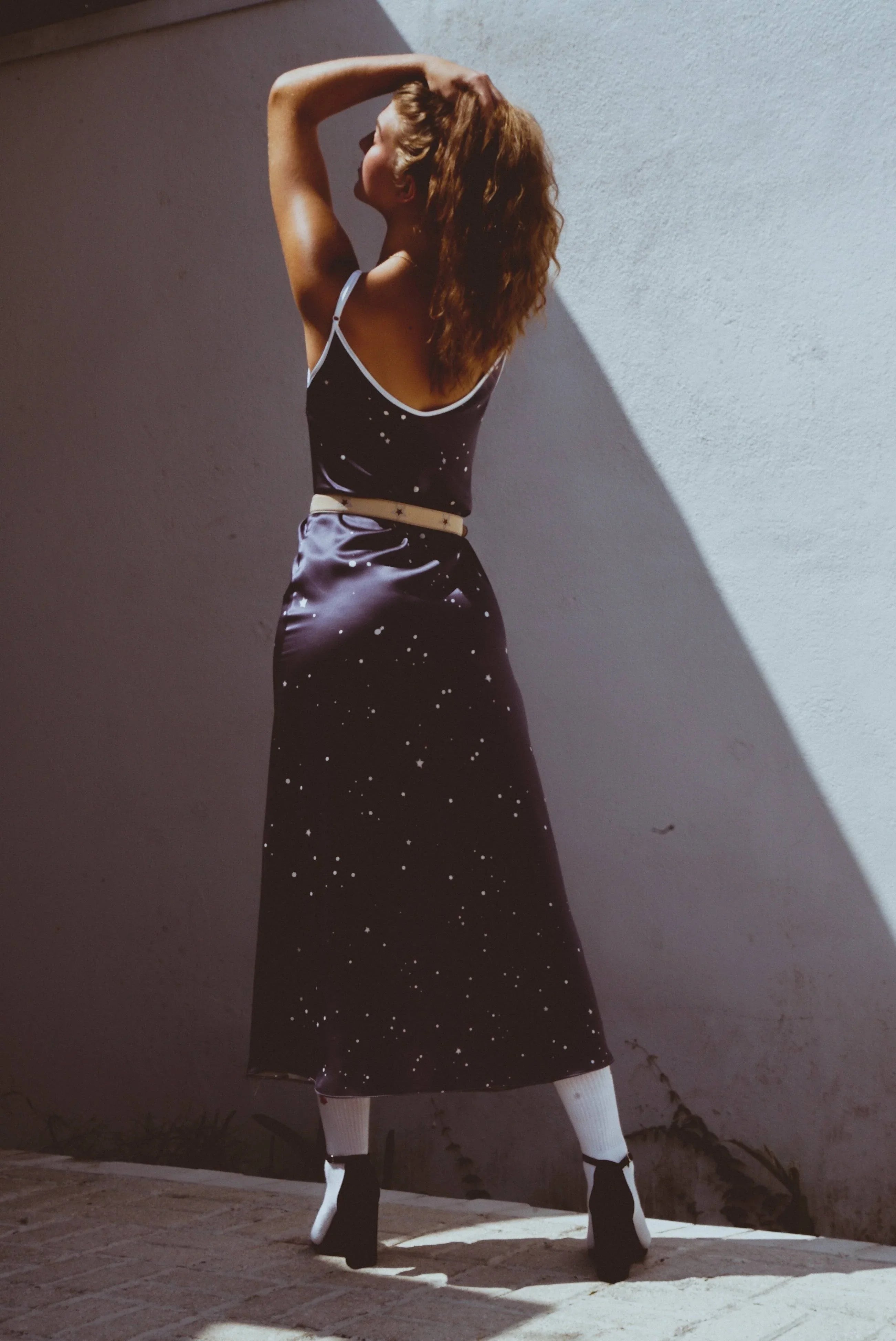 Woman in black starry dress, white belt and socks, posing against light blue wall in sunlight