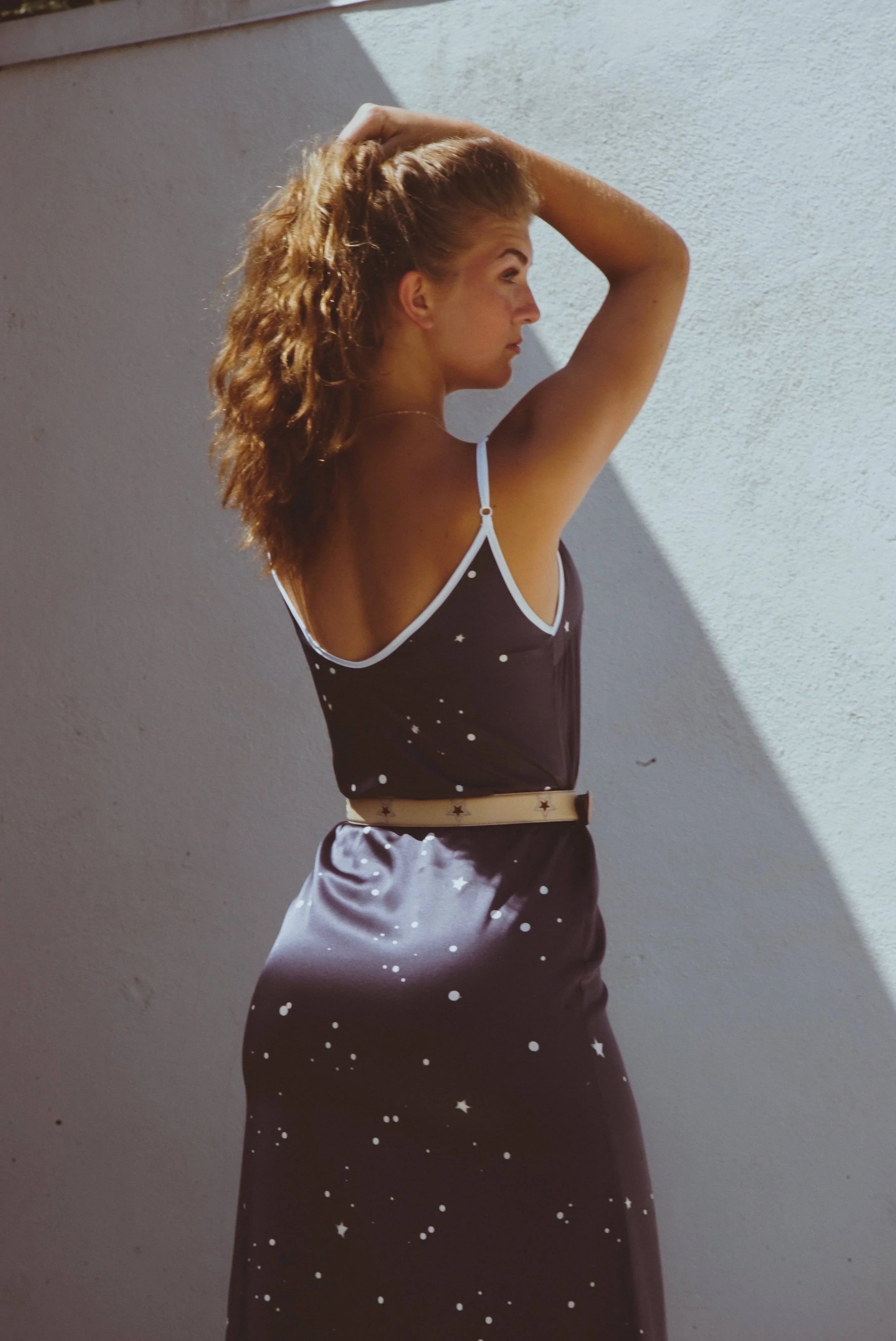 Young woman with curly hair wearing a dark starry print dress and beige belt, posing against a light textured wall