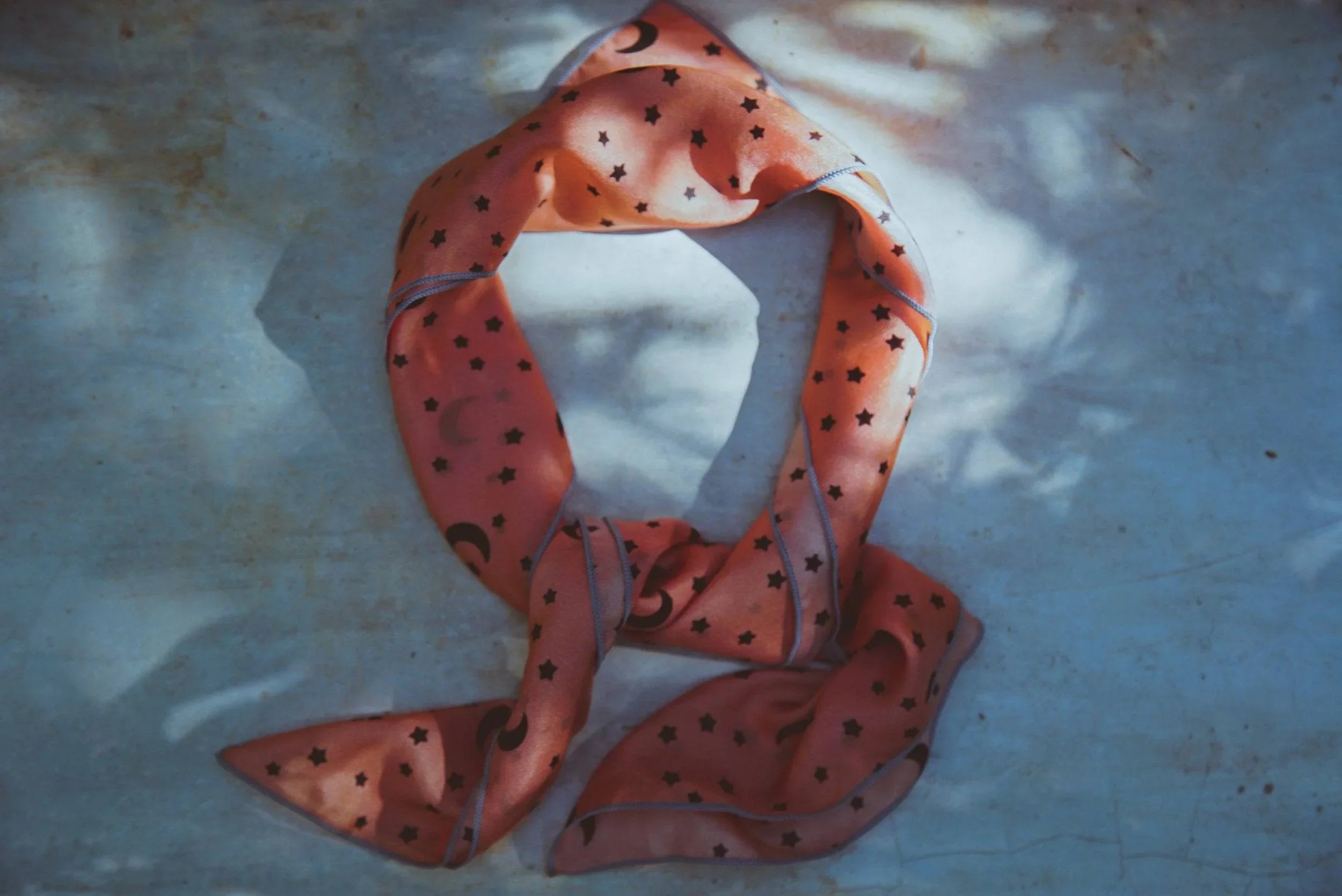 Orange scarf with moon and star prints on textured blue-gray surface under dappled light