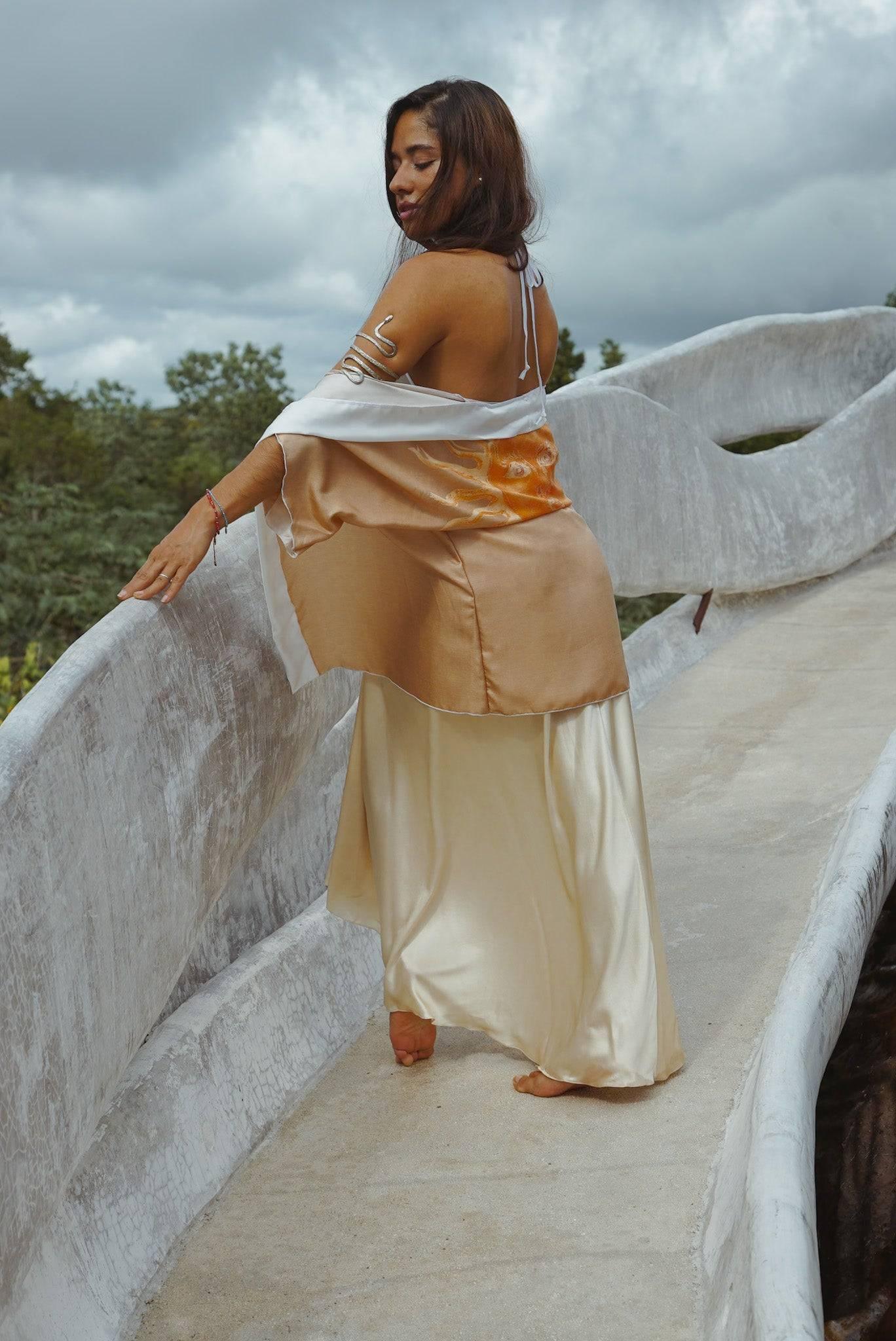 Woman in flowing satin dress and shawl standing barefoot on curved textured bridge under cloudy sky