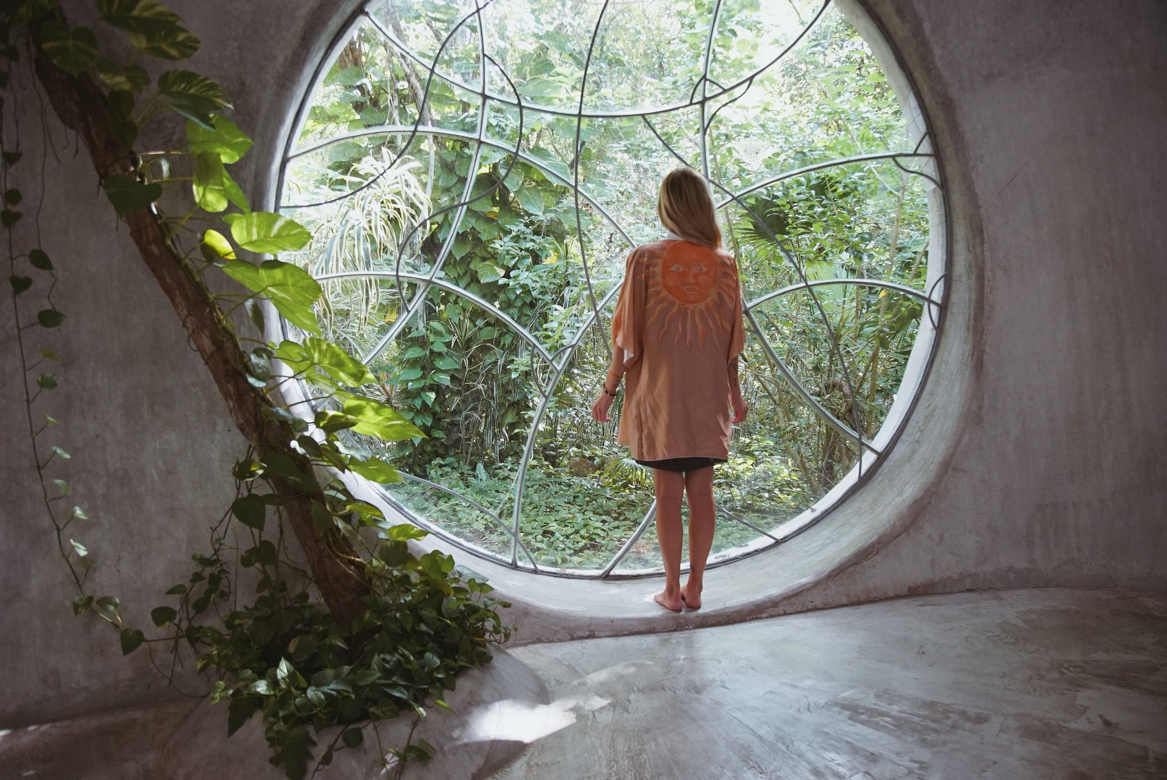 Woman wearing sun print jacket standing barefoot by large round window with jungle greenery outside