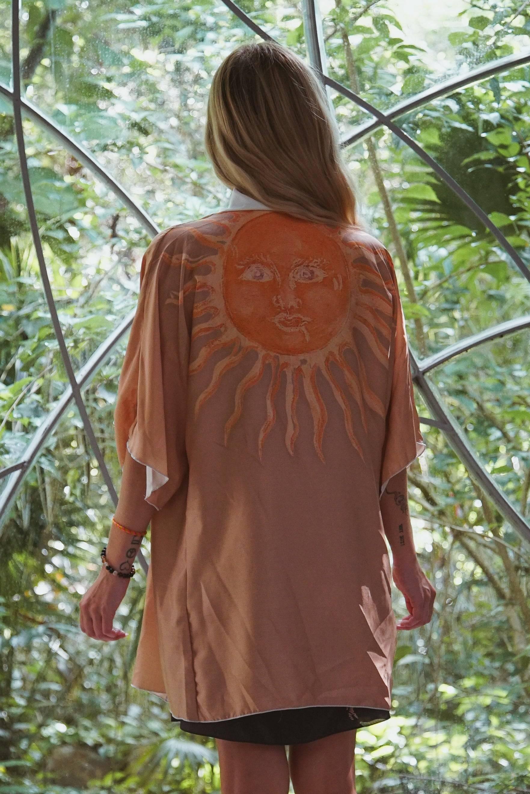 Woman wearing a beige kimono with an orange sun face design, standing in a lush green garden