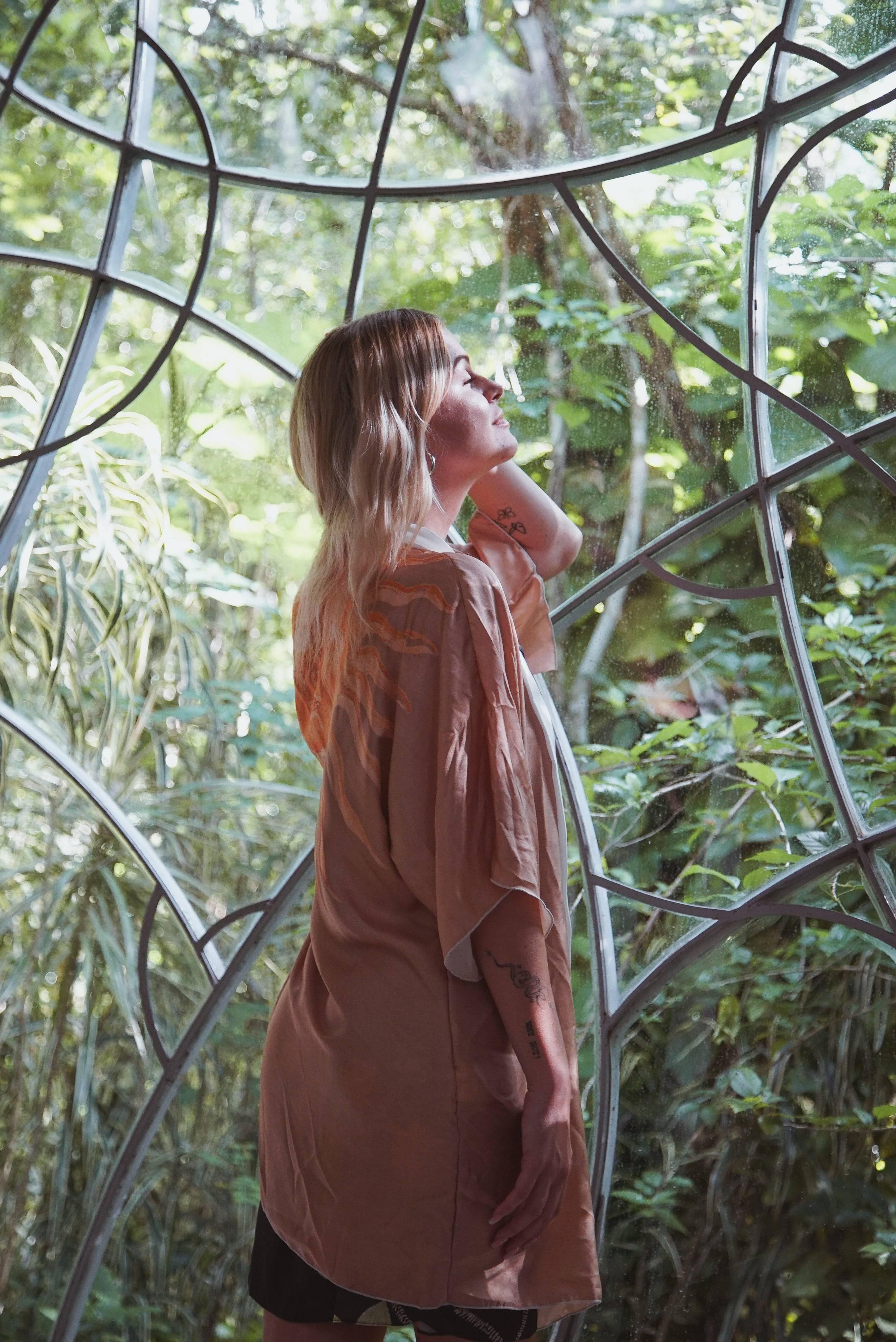 Blonde woman with tattoos wearing peach kimono by large geometric window with lush green plants outside