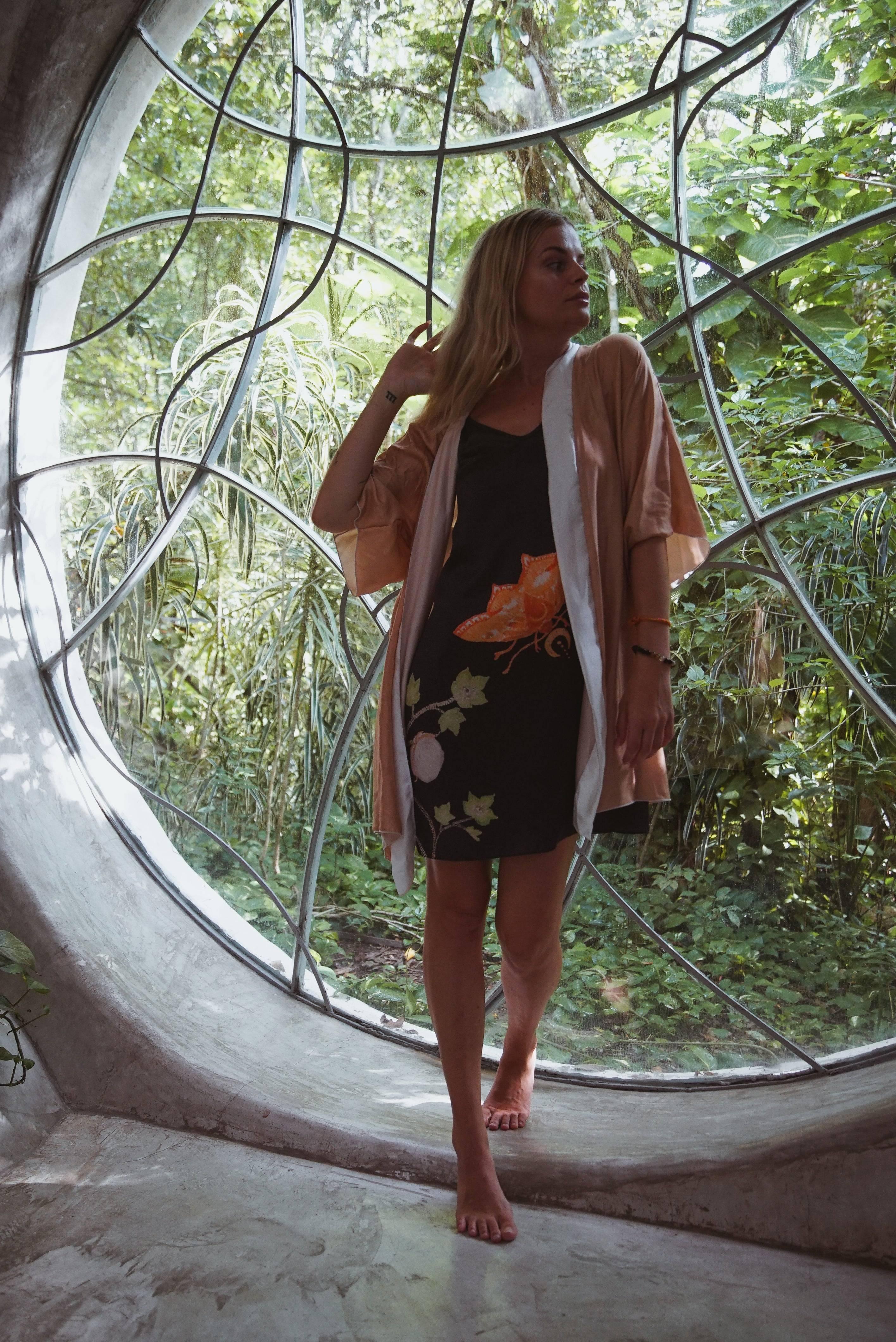 Woman in floral dress and peach kimono standing barefoot by artistic round window with jungle view