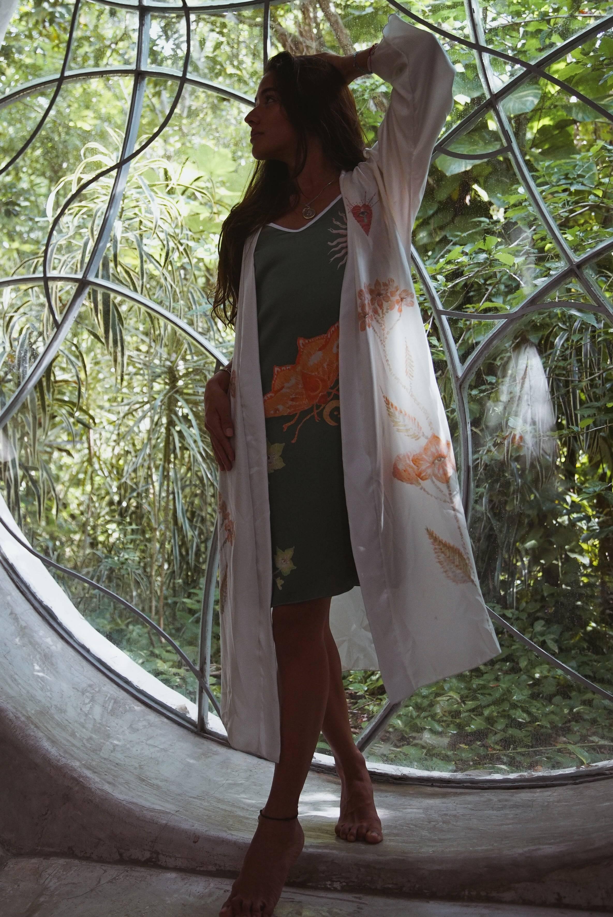 Woman in floral kimono and green dress posing barefoot in a circular window with lush greenery outside