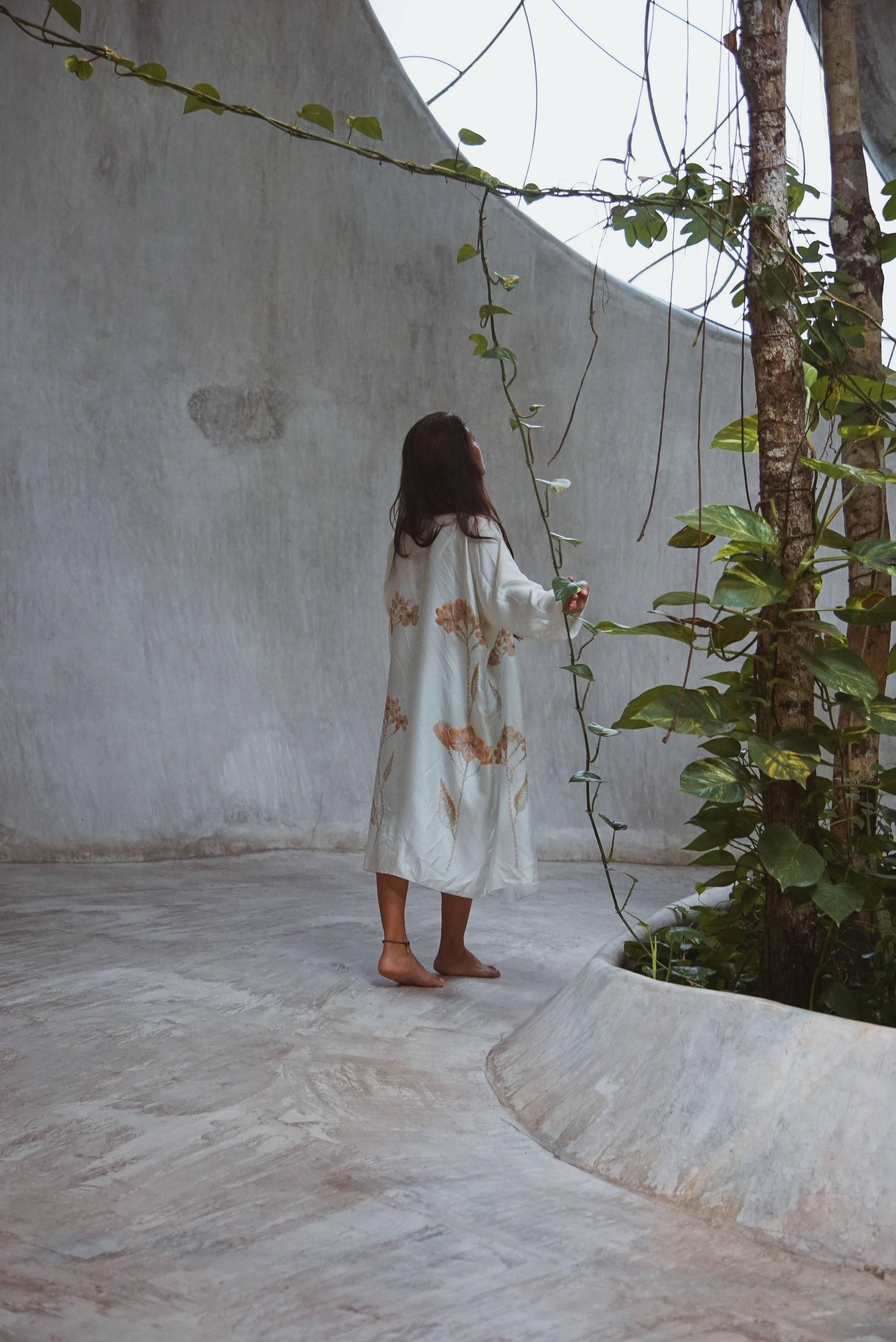 Woman in a white floral dress barefoot in a minimalistic indoor garden with concrete floor and plants