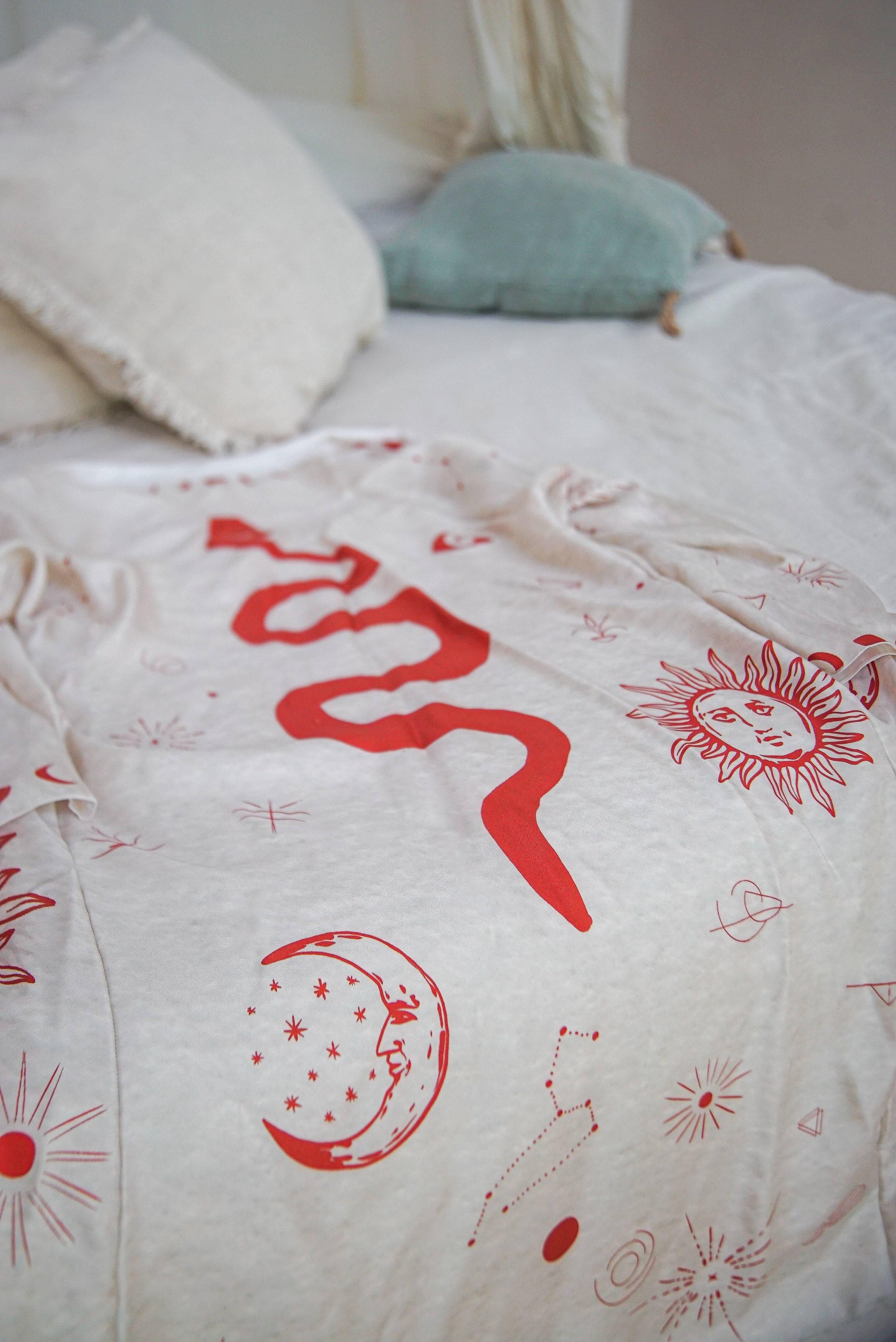 White t-shirt with red celestial and serpent designs laid on white bed with pillows