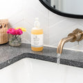Mama Suds hand soap on granite bathroom counter by running brass faucet, flowers in vase