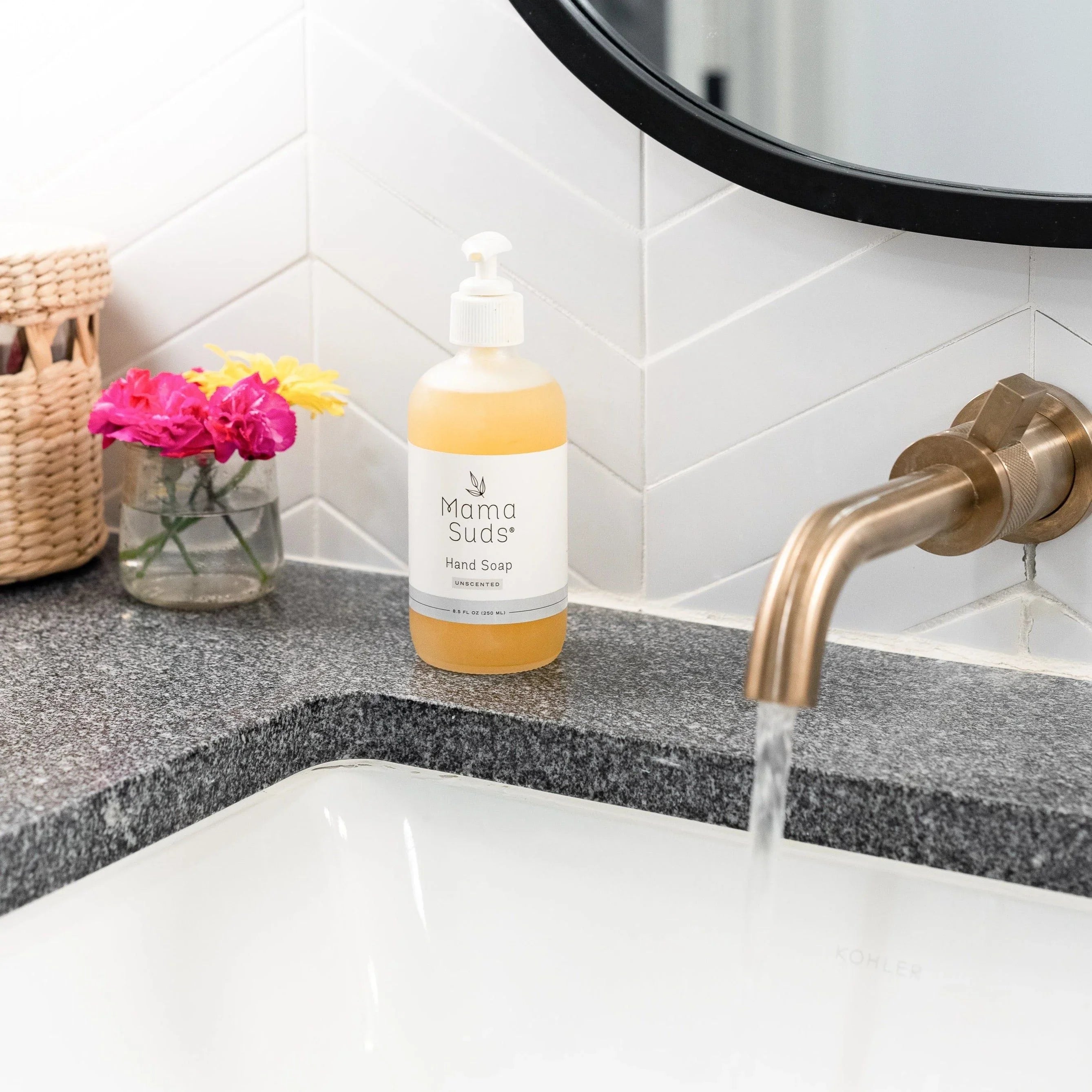 Mama Suds hand soap on granite bathroom counter by running brass faucet, flowers in vase