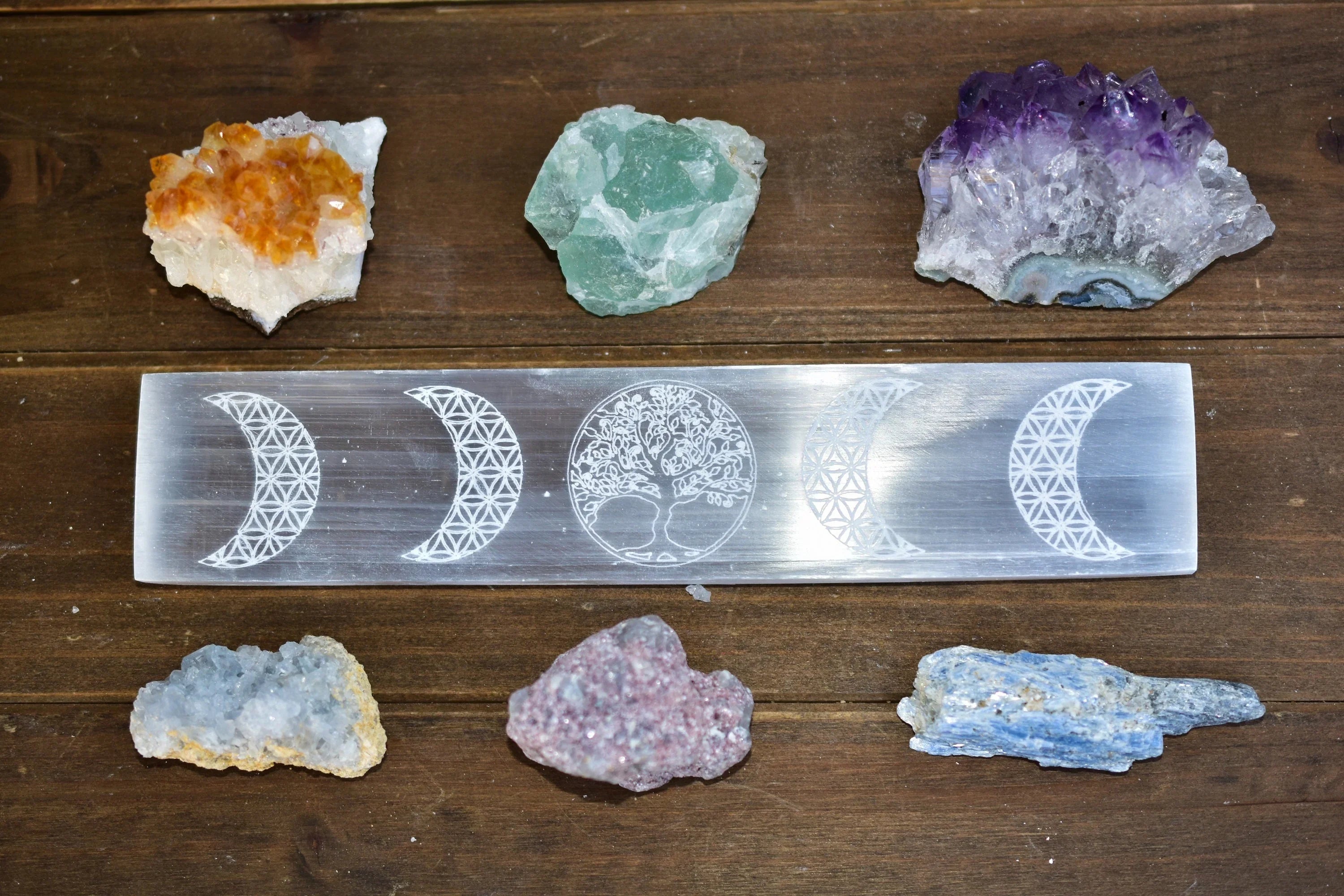 Selenite crystal engraved with crescent moons and tree of life surrounded by six colorful mineral stones on wooden surface