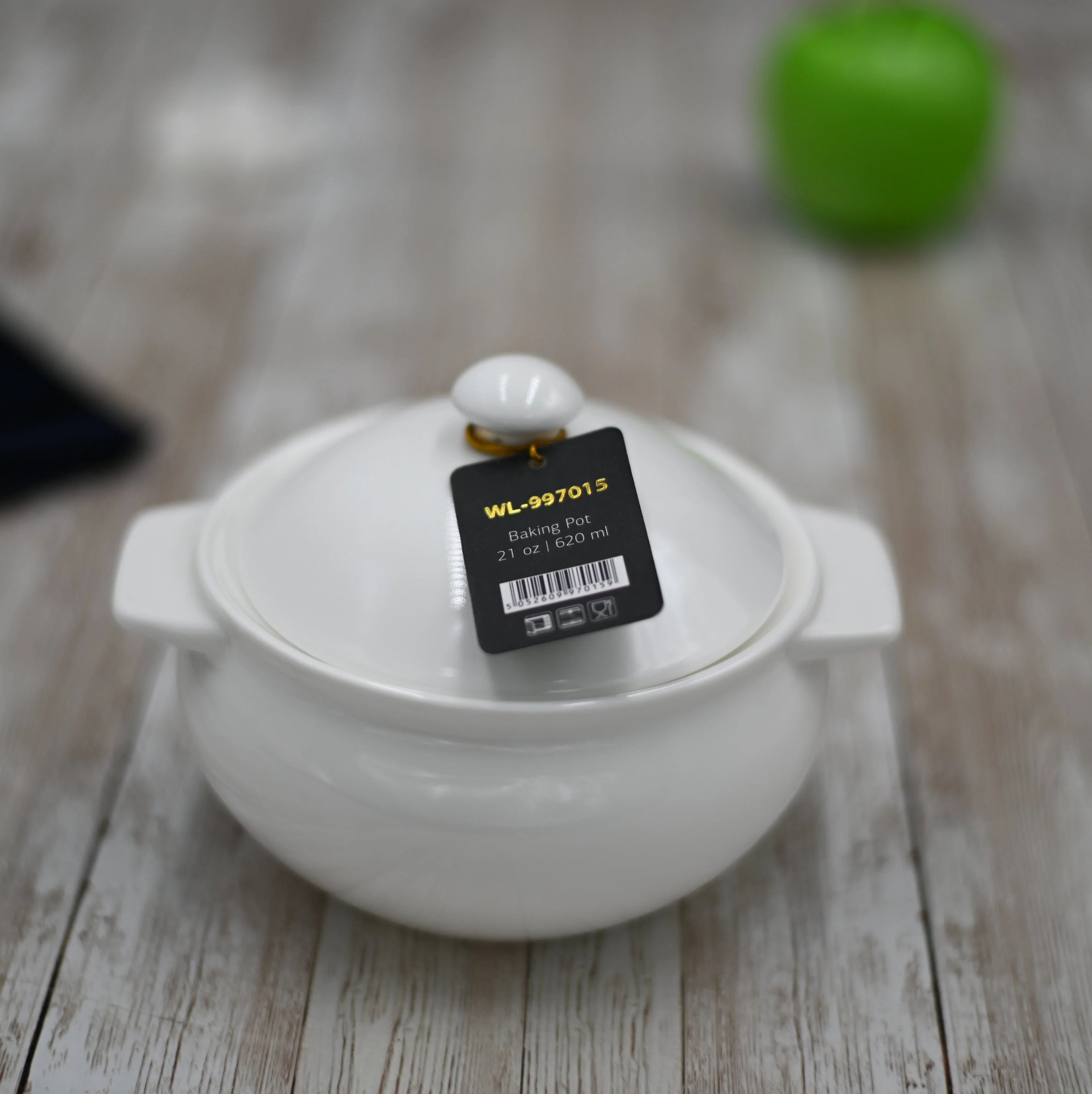 White ceramic baking pot with lid and tag, 21 oz, on wooden surface with blurred green apple