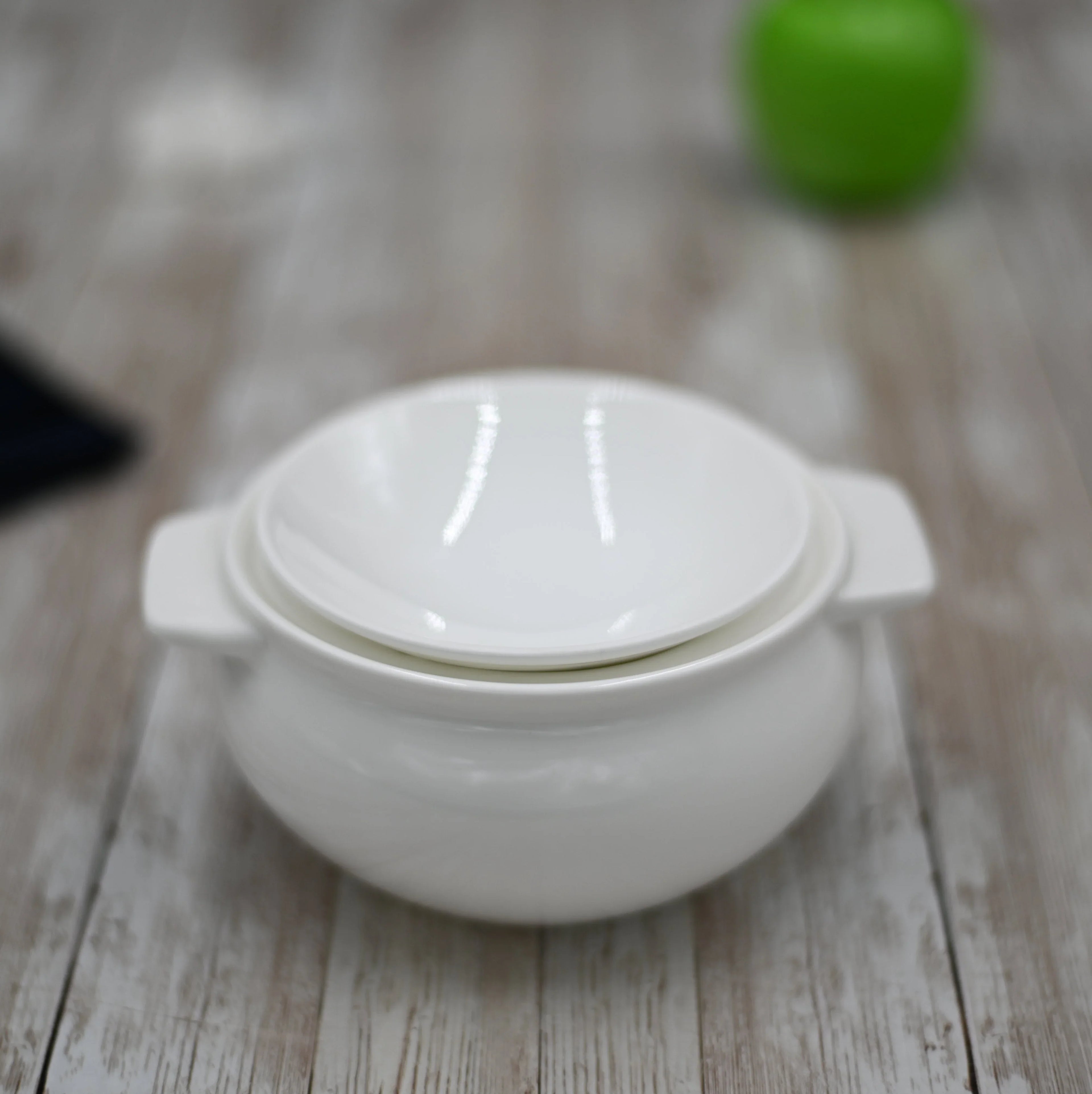 White ceramic soup bowl with lid on rustic wooden table, green blurred object background