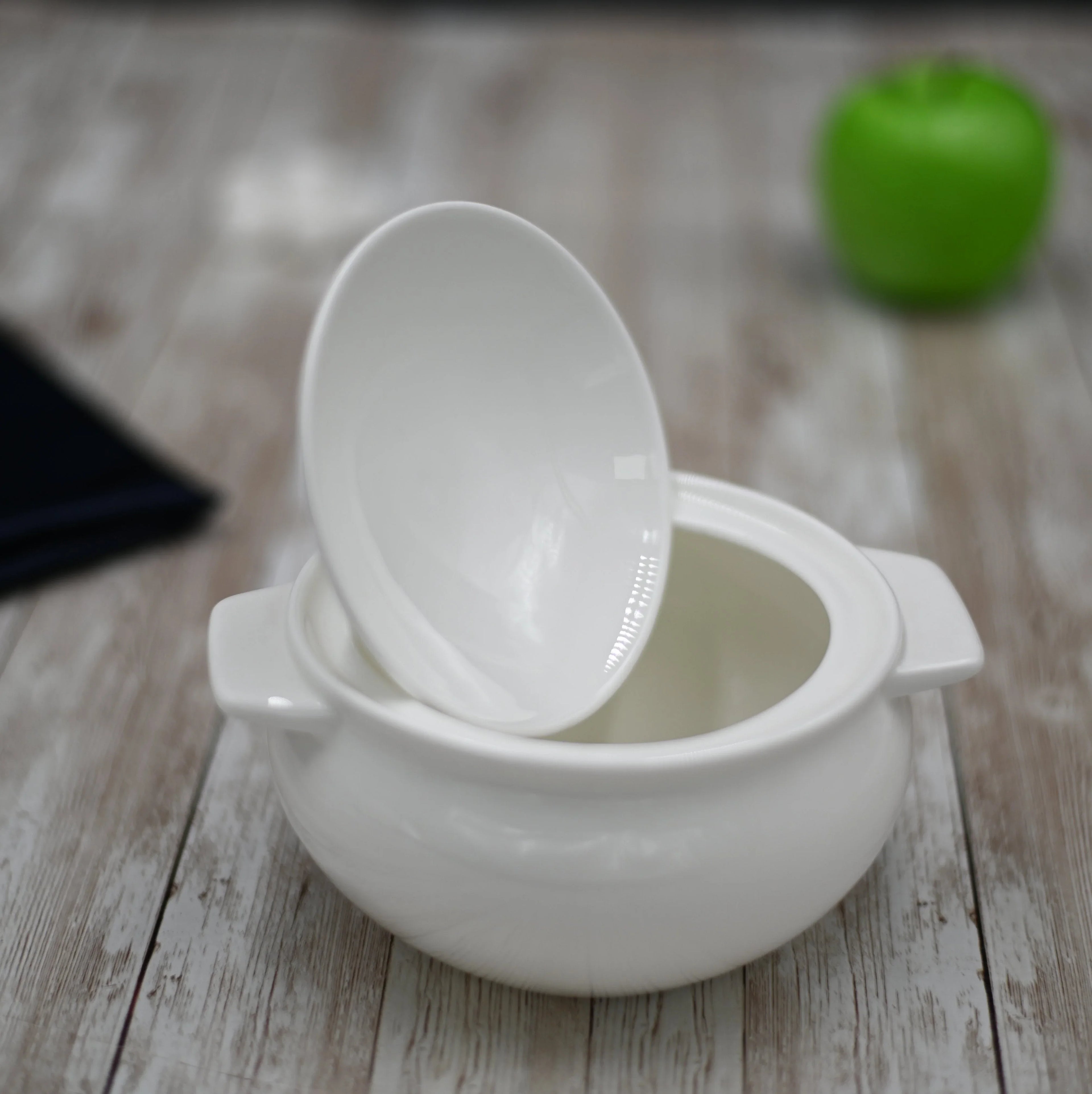 White ceramic soup tureen with lid on wooden table and green apple in background