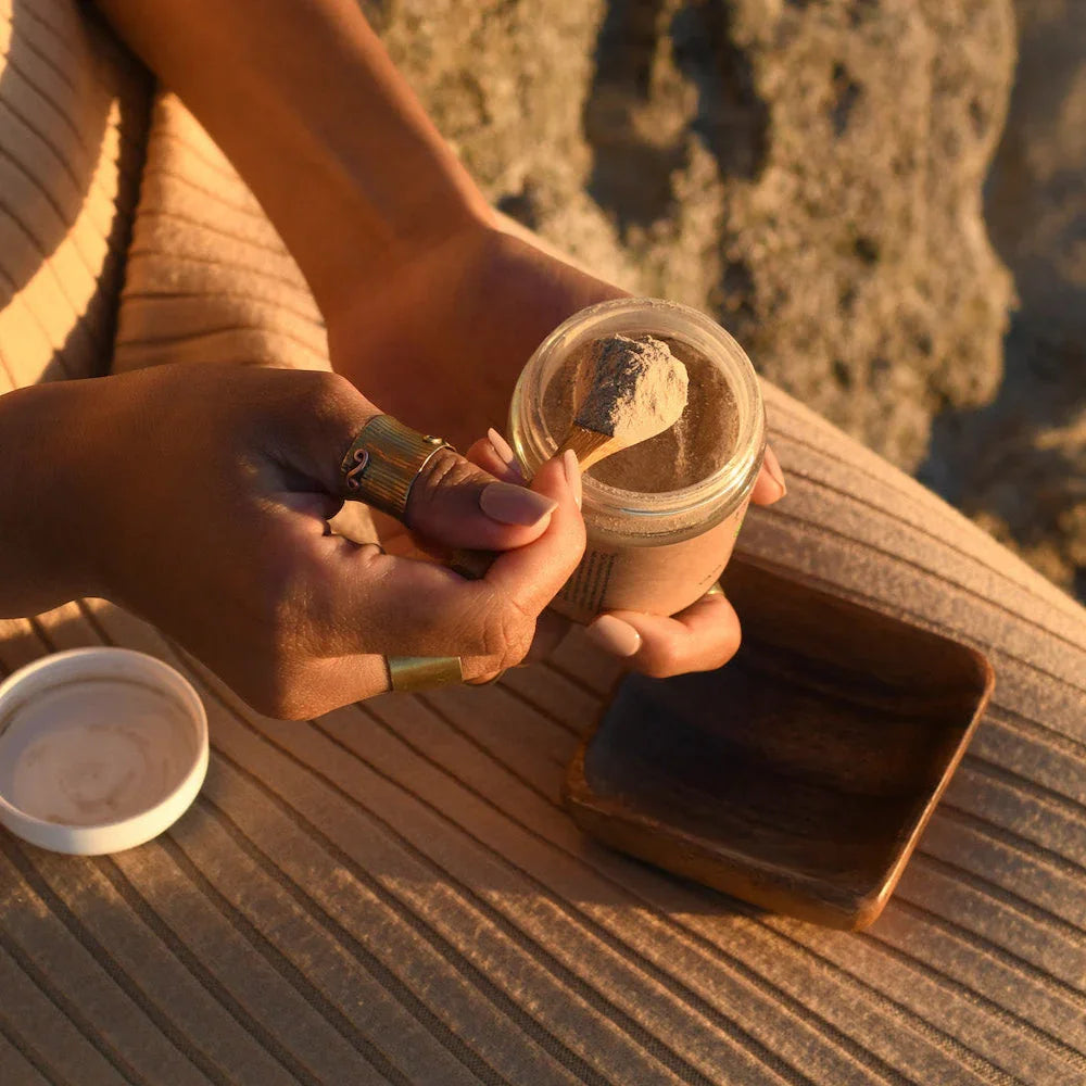 Hands holding a jar of brown clay mask powder with a wooden spoon, on a ribbed fabric and wooden bowl
