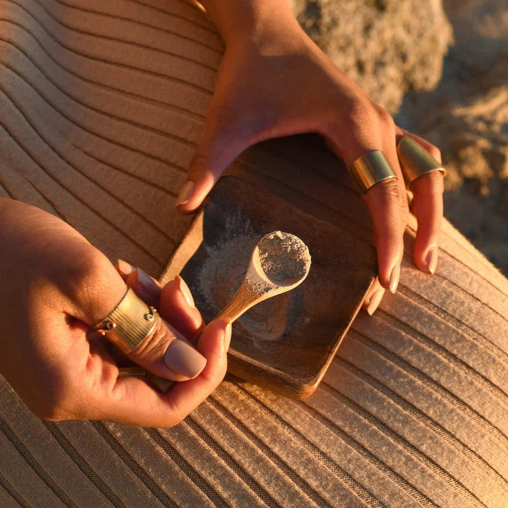 Hands with gold rings holding a wooden bowl and spoon with powdered clay on a textured fabric surface