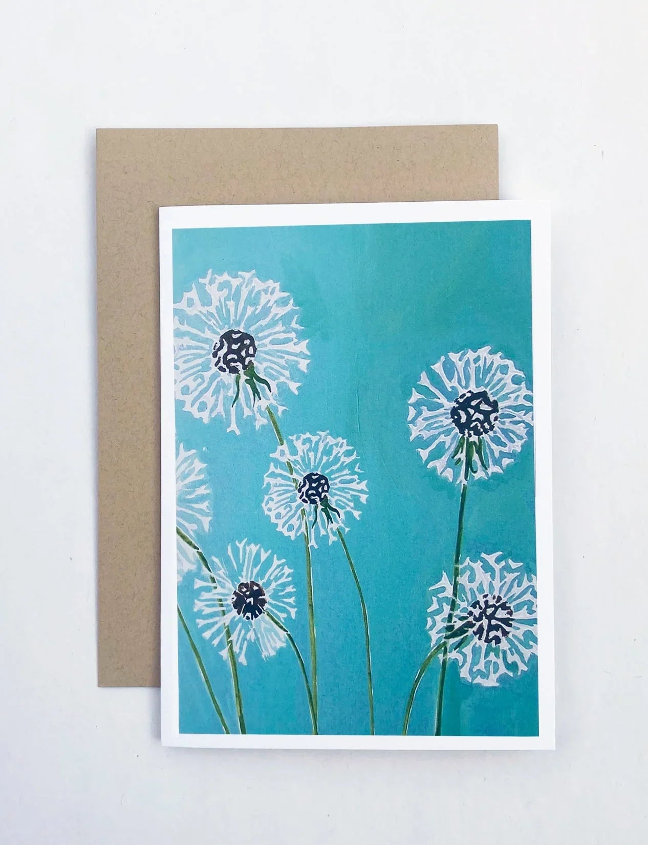 Notecard with white dandelion illustrations on a turquoise background and a brown envelope