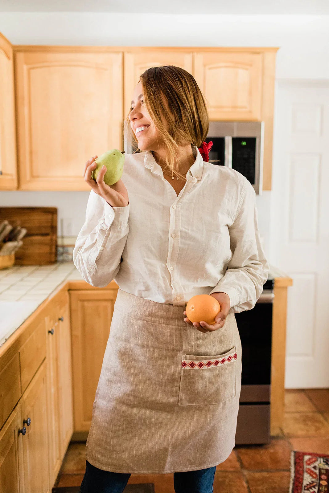 Smiling woman in artisan apron holding fruit, standing in cozy kitchen at Qu!rk Boutique.