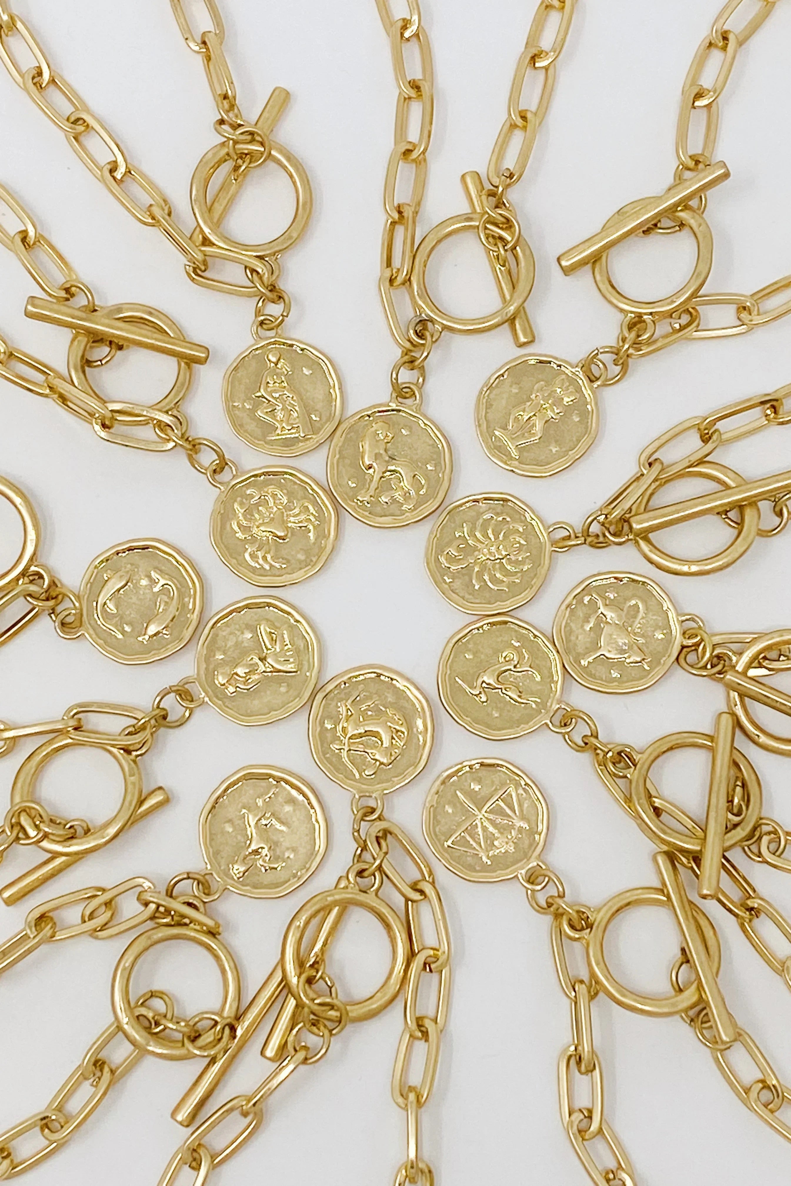 Gold zodiac sign necklaces with toggle clasps arranged in a circular pattern on white background