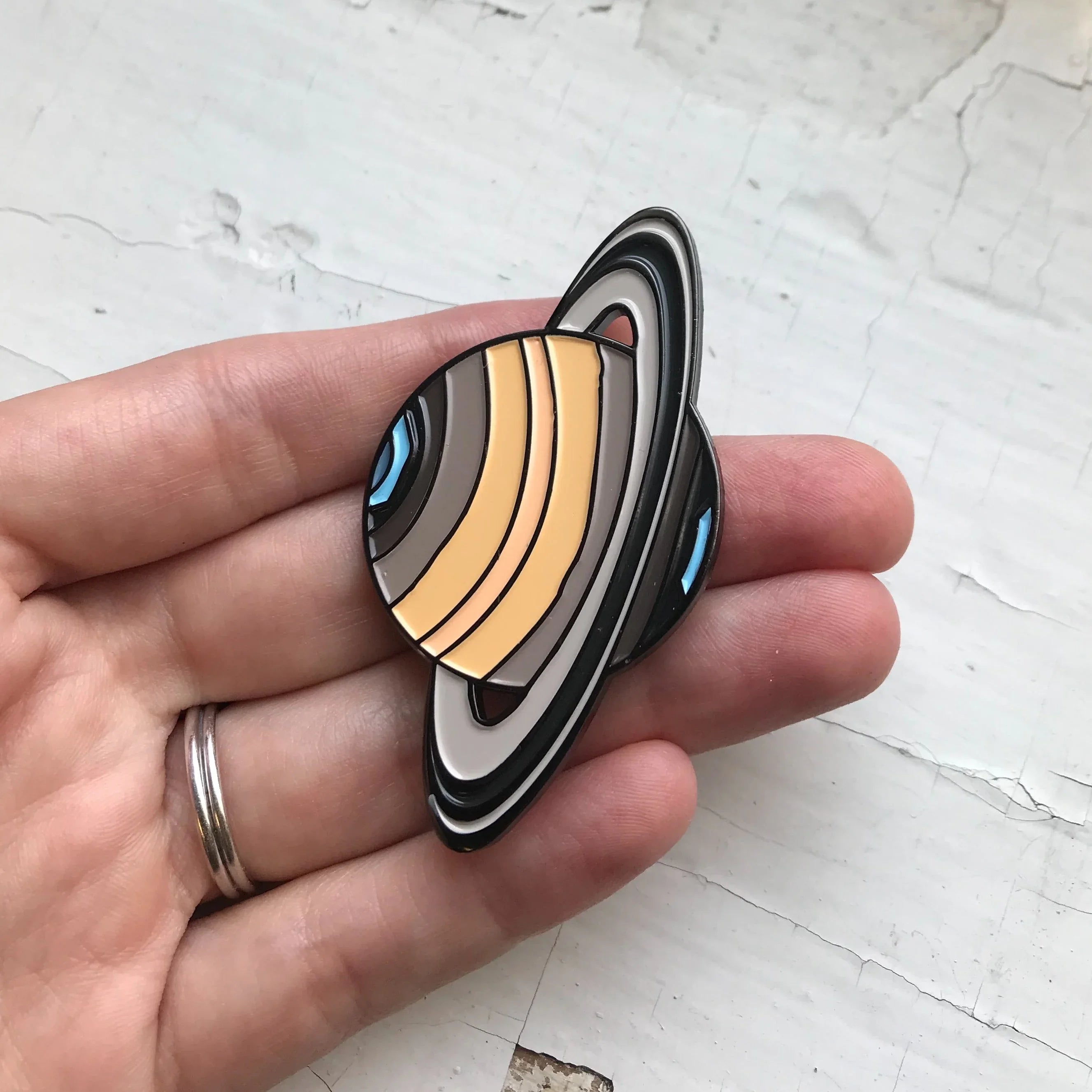 Hand holding Saturn planet enamel pin with cosmic design from Qu!rk Boutique