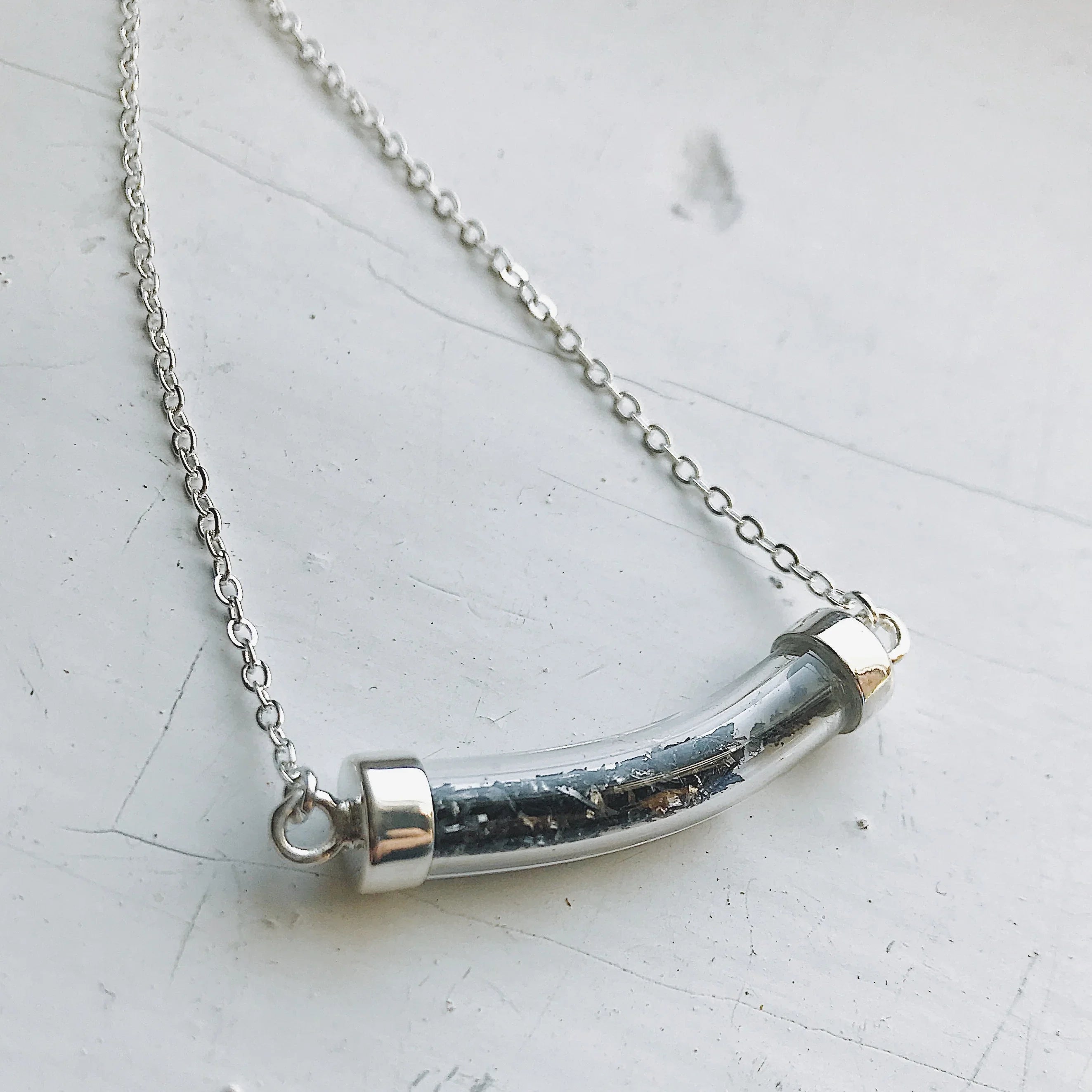 Silver necklace with a curved glass pendant filled with small black and metallic flakes on white surface