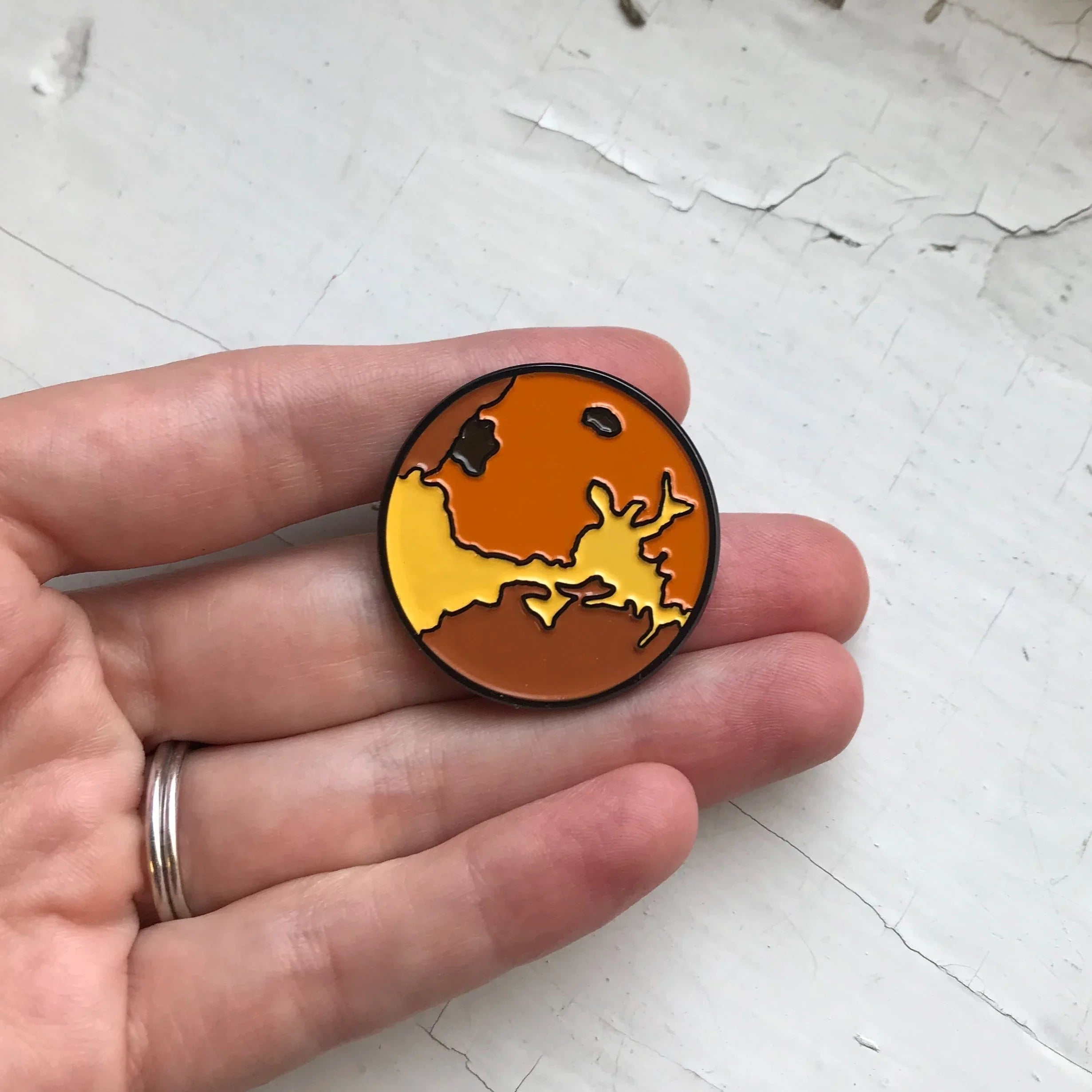 Hand holding a round pin with abstract orange and yellow map design on a white textured surface