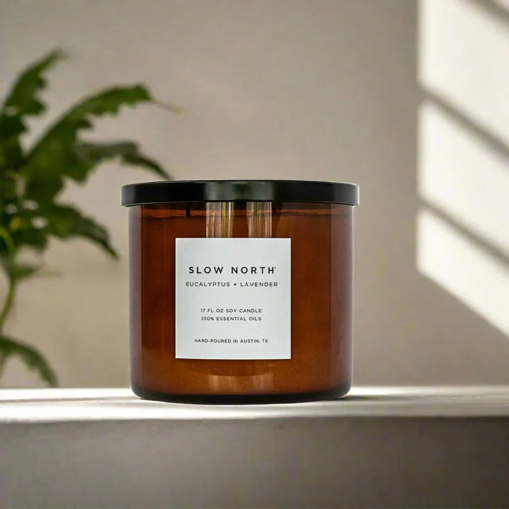 Slow North eucalyptus and lavender soy candle in amber glass jar on white surface with plant background