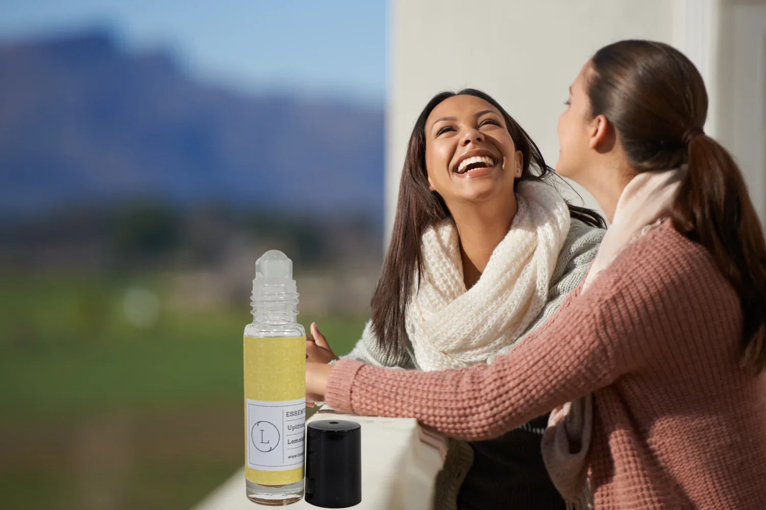 Two women wearing scarves laughing outdoors near a bottle of uplifting lemon essential oil