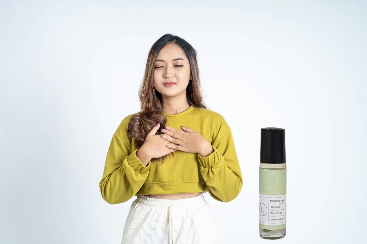 Young woman in green sweater with eyes closed holding hands to chest next to tea tree essential oil bottle