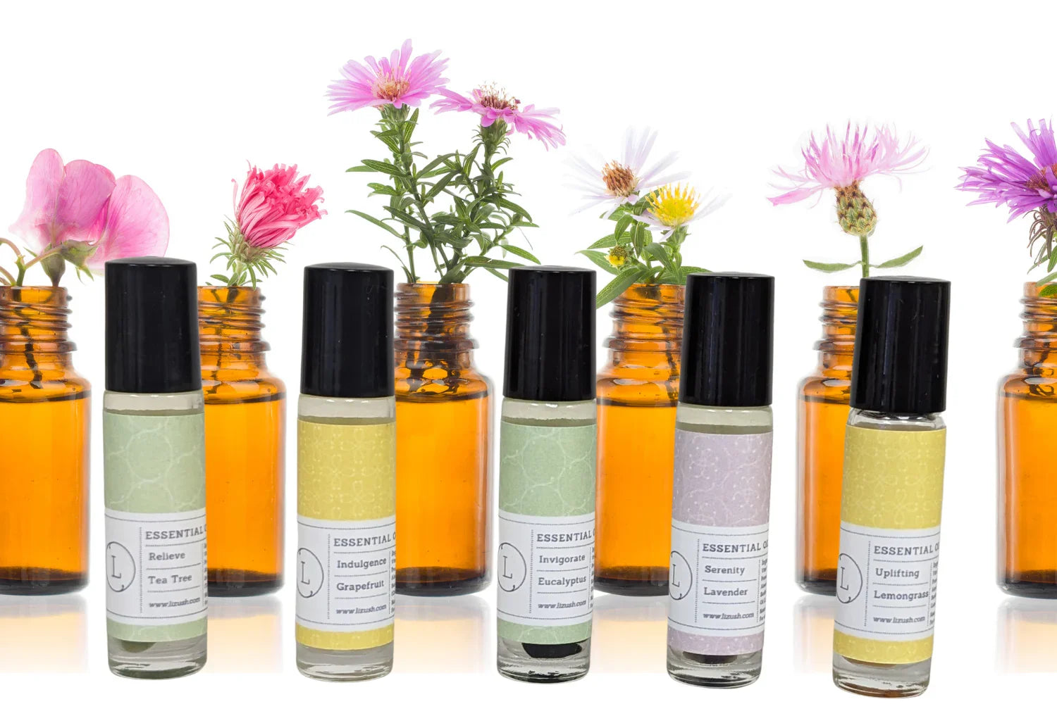 Roll-on essential oils with black caps and pastel labels in front of amber bottles holding pink, white, and purple flowers