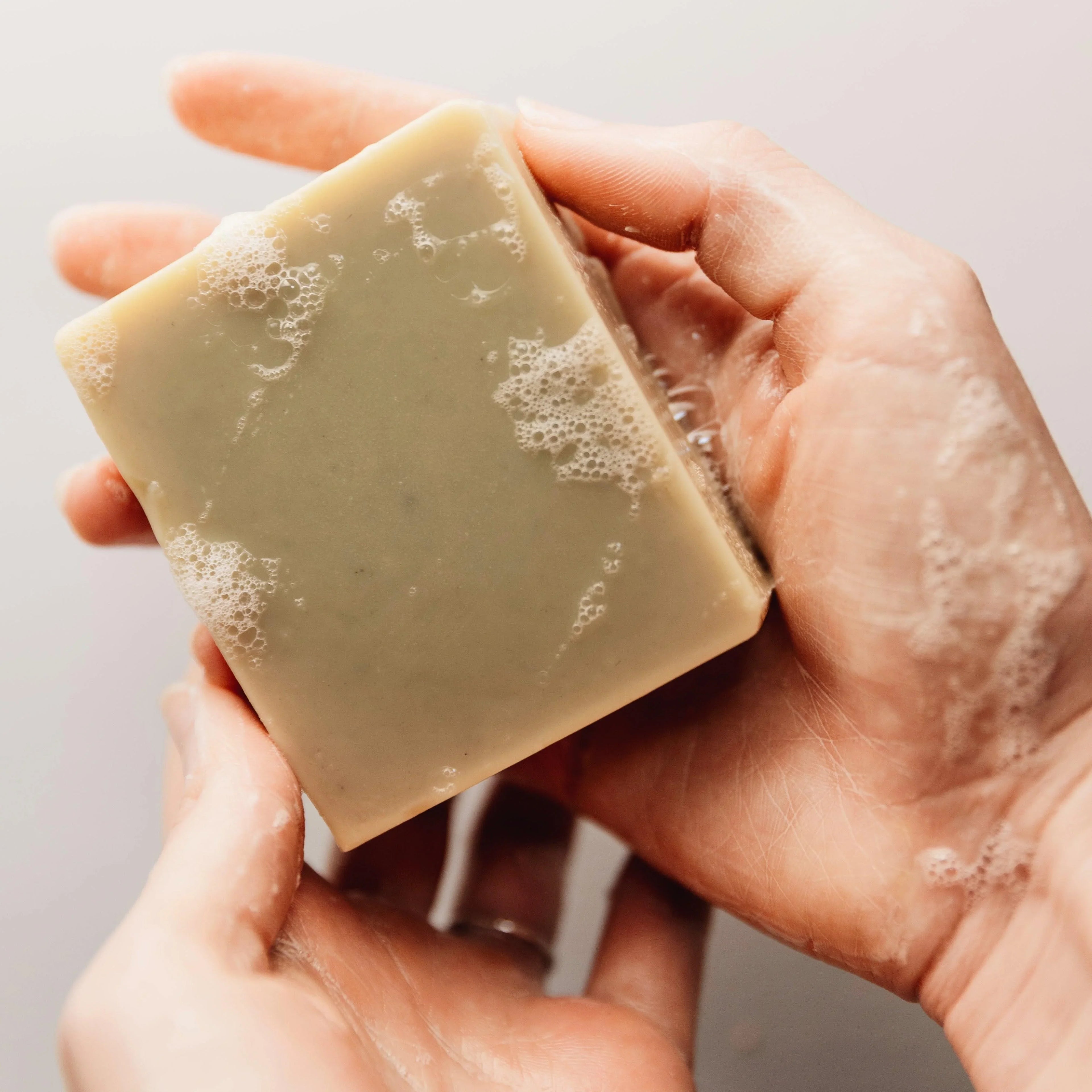 Hands holding a lathered beige natural bar soap with foam bubbles