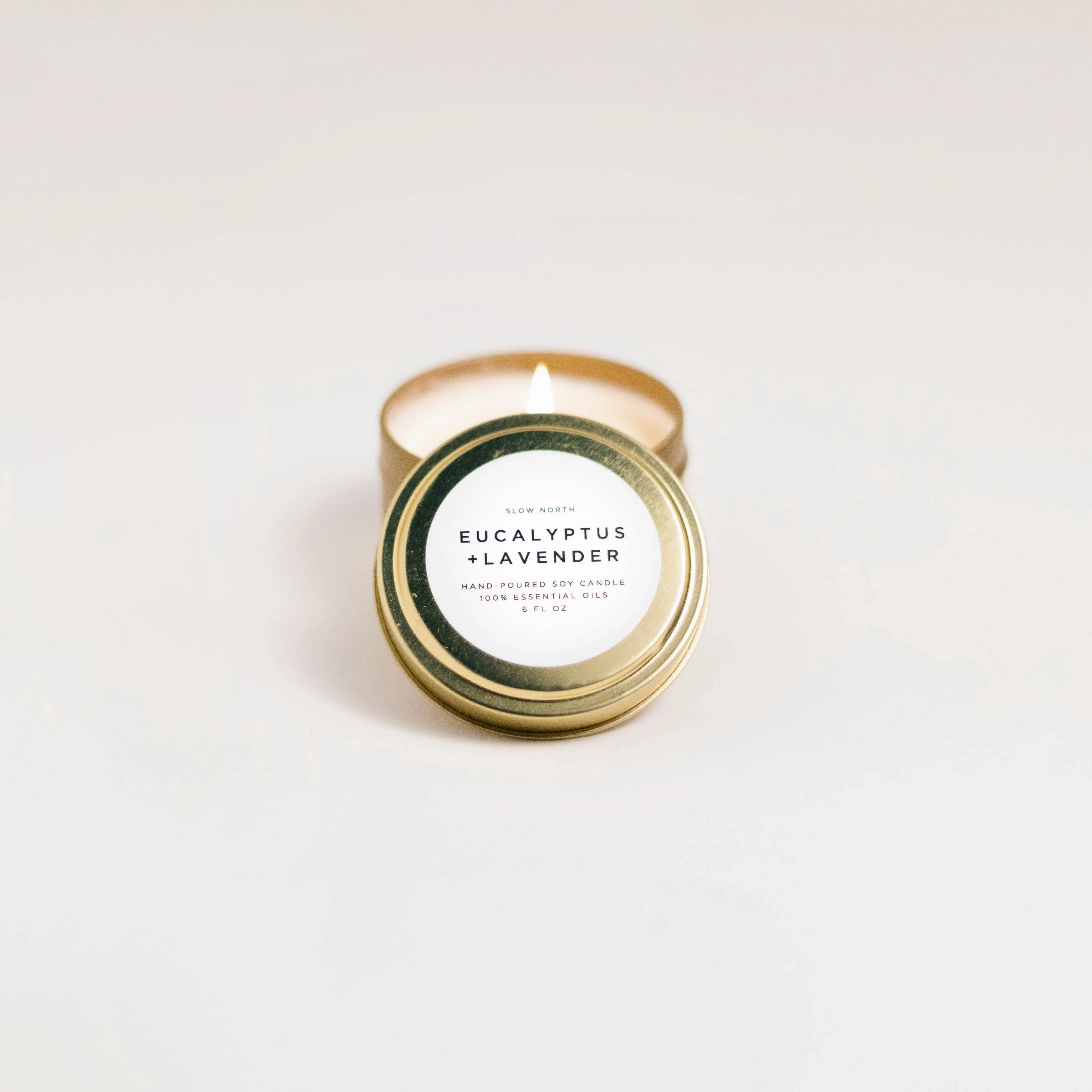 Lit eucalyptus and lavender scented soy candle in gold travel tin on white background