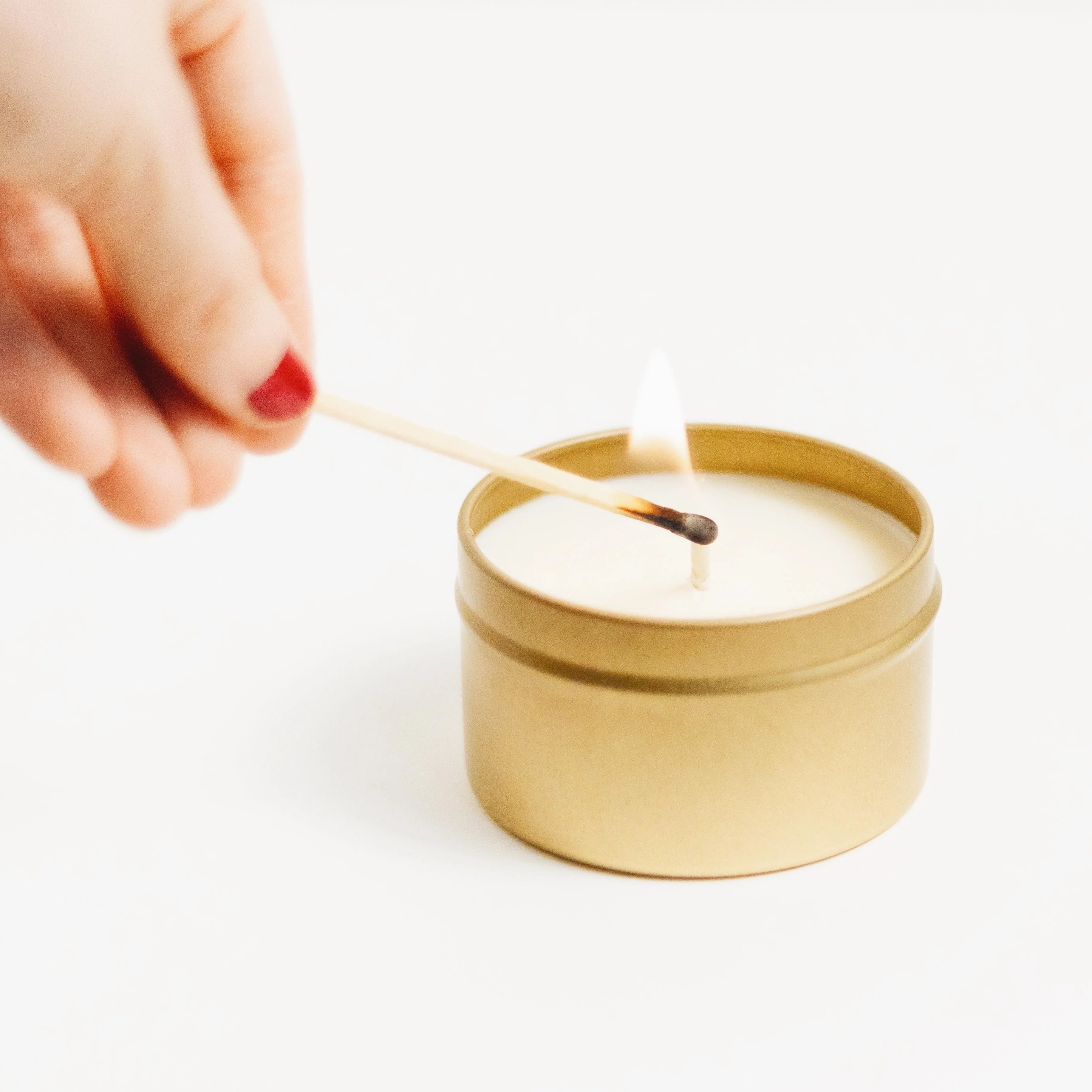 Hand lighting a gold travel tin candle with a matchstick on white background