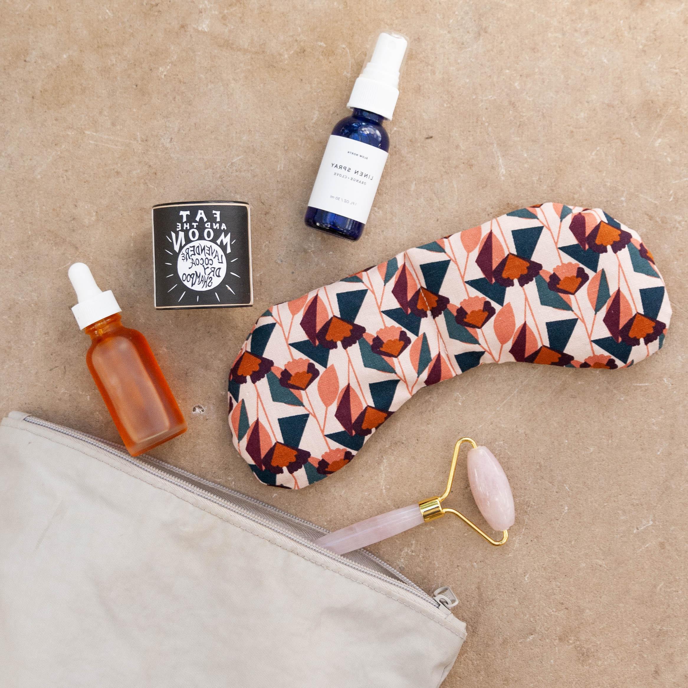 Floral patterned eye mask, face roller, amber dropper bottle, blue spray bottle, and cream on beige background