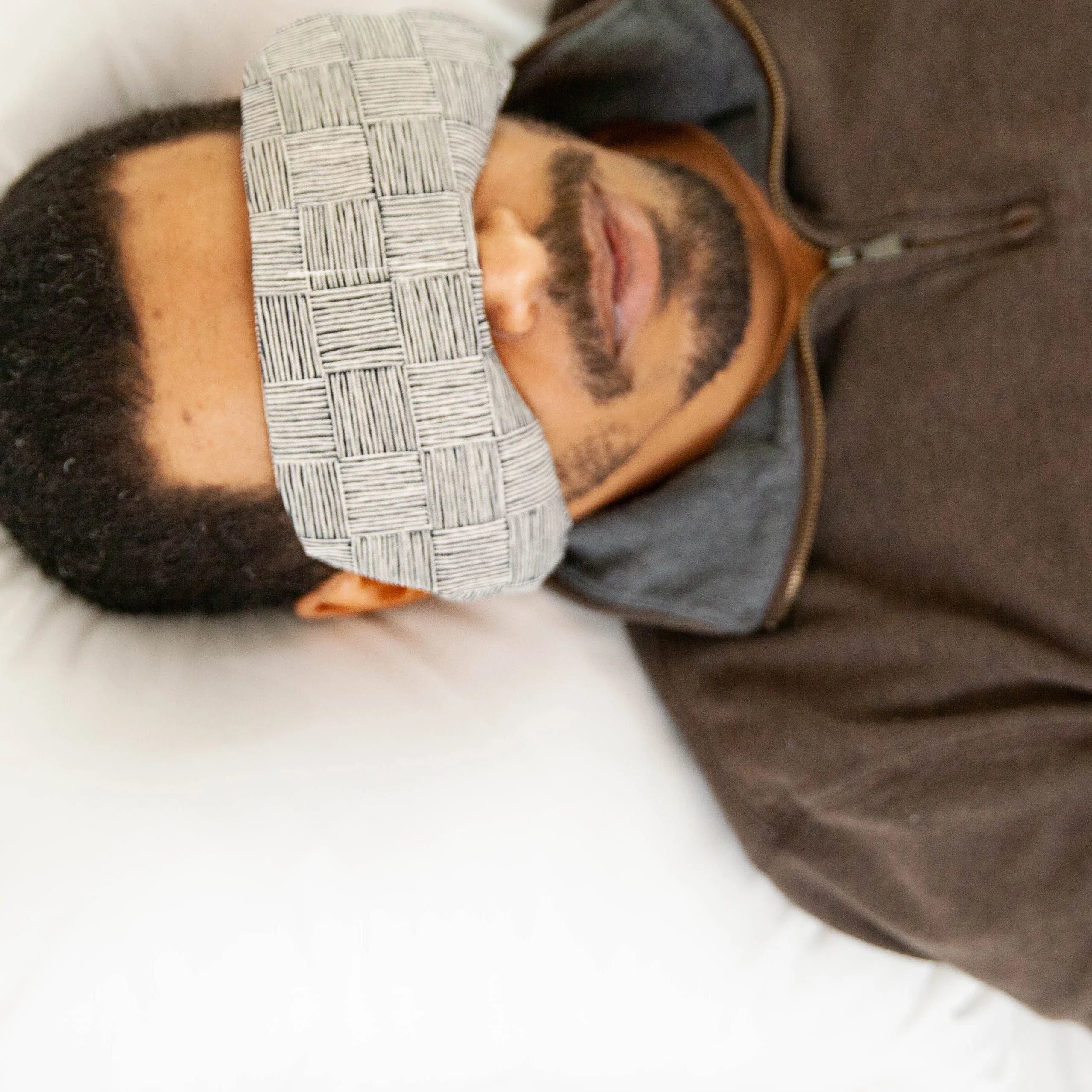 Man resting with woven patterned eye mask, cozy in brown zip sweater on white bedding