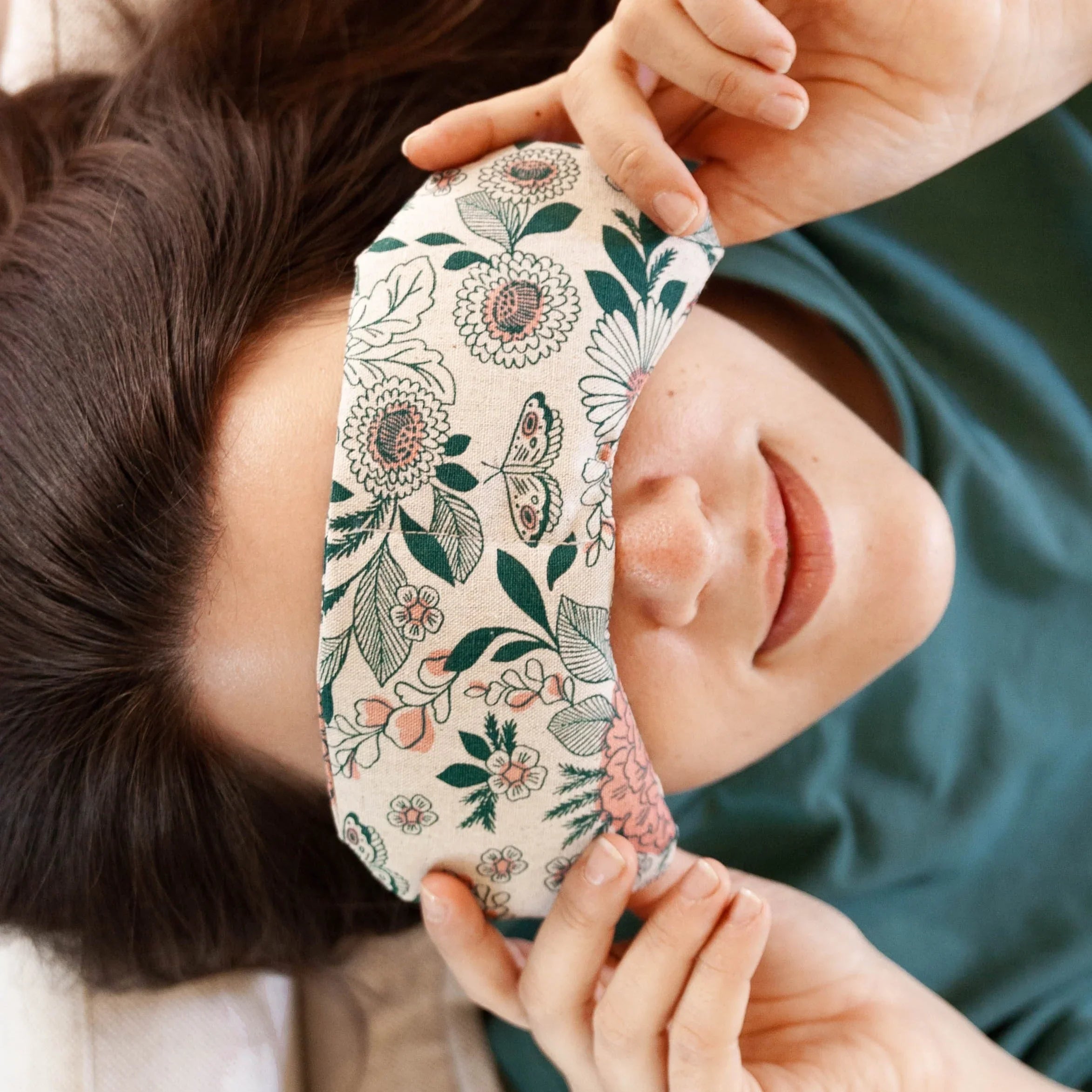 Close-up of woman lying down with floral patterned eye mask on face, hands adjusting it, wearing green shirt