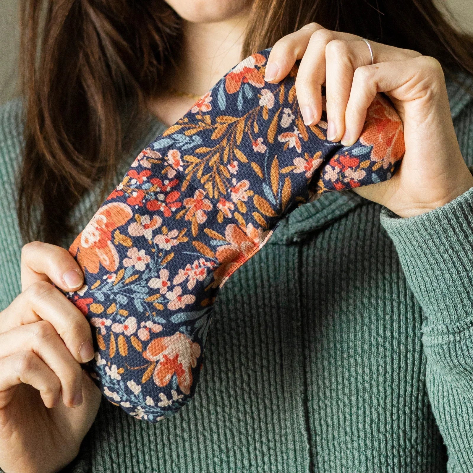 Person holding a floral patterned eye mask with pink, orange, and blue flowers, wearing a green textured sweater