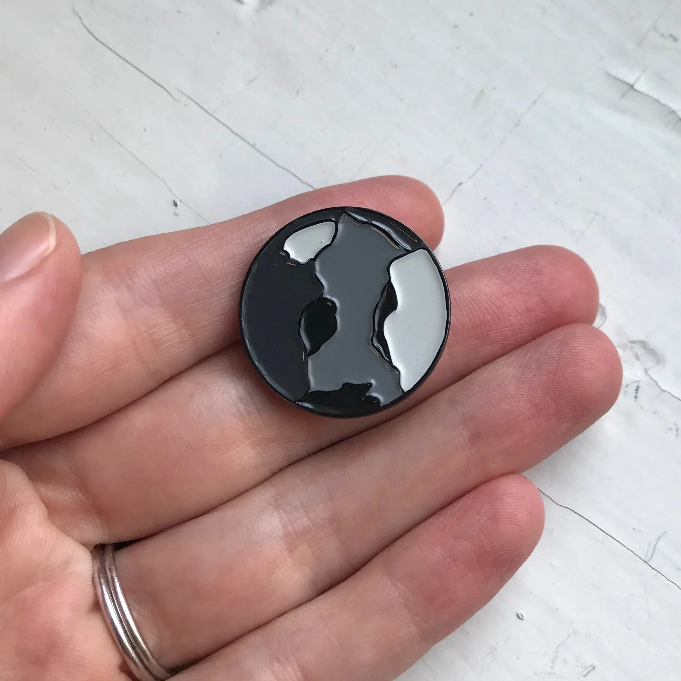 Hand holding a round enamel pin shaped like a black and gray abstract globe
