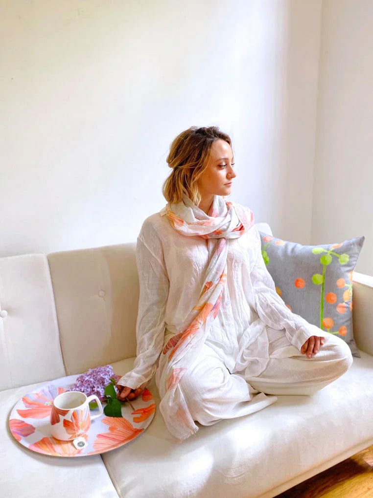 Woman in white outfit and floral scarf sitting cross-legged on cream sofa with floral tray and mug in cozy room