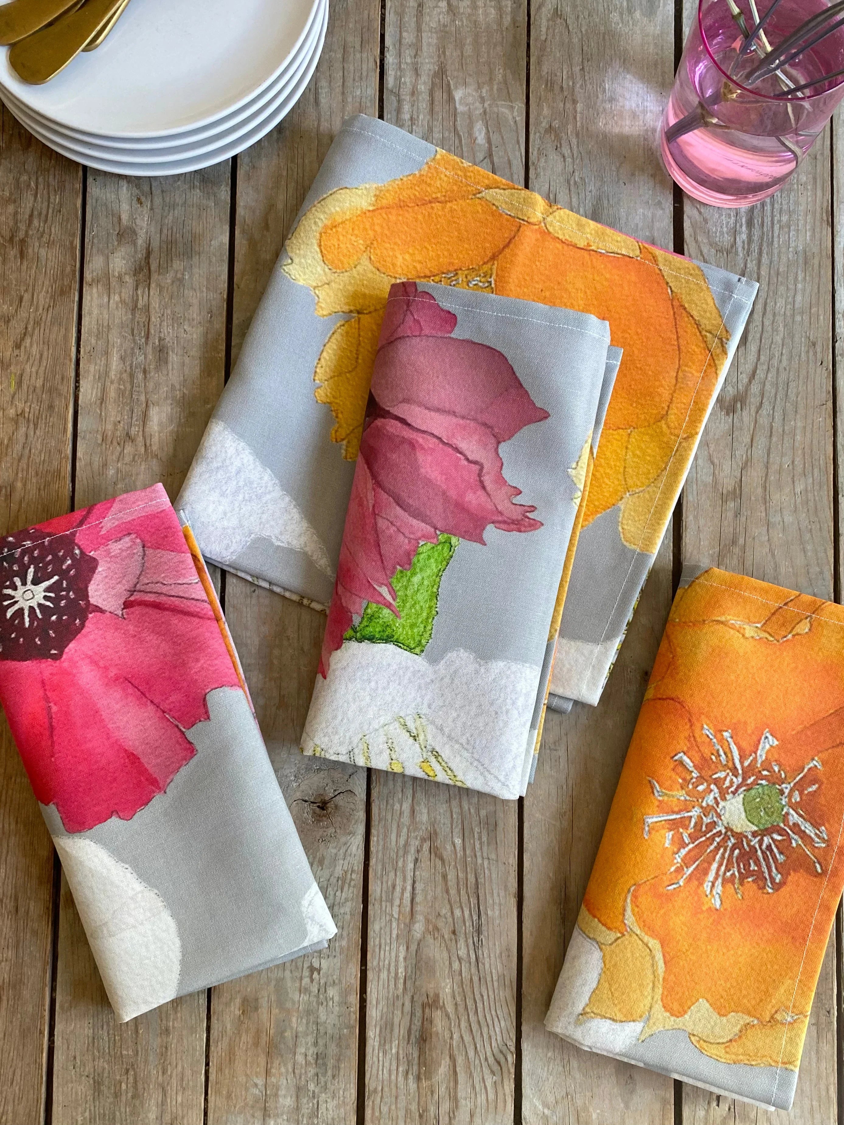 Colorful floral fabric napkins in pink, orange, and yellow on rustic wooden tabletop with stacked white plates and pink glass