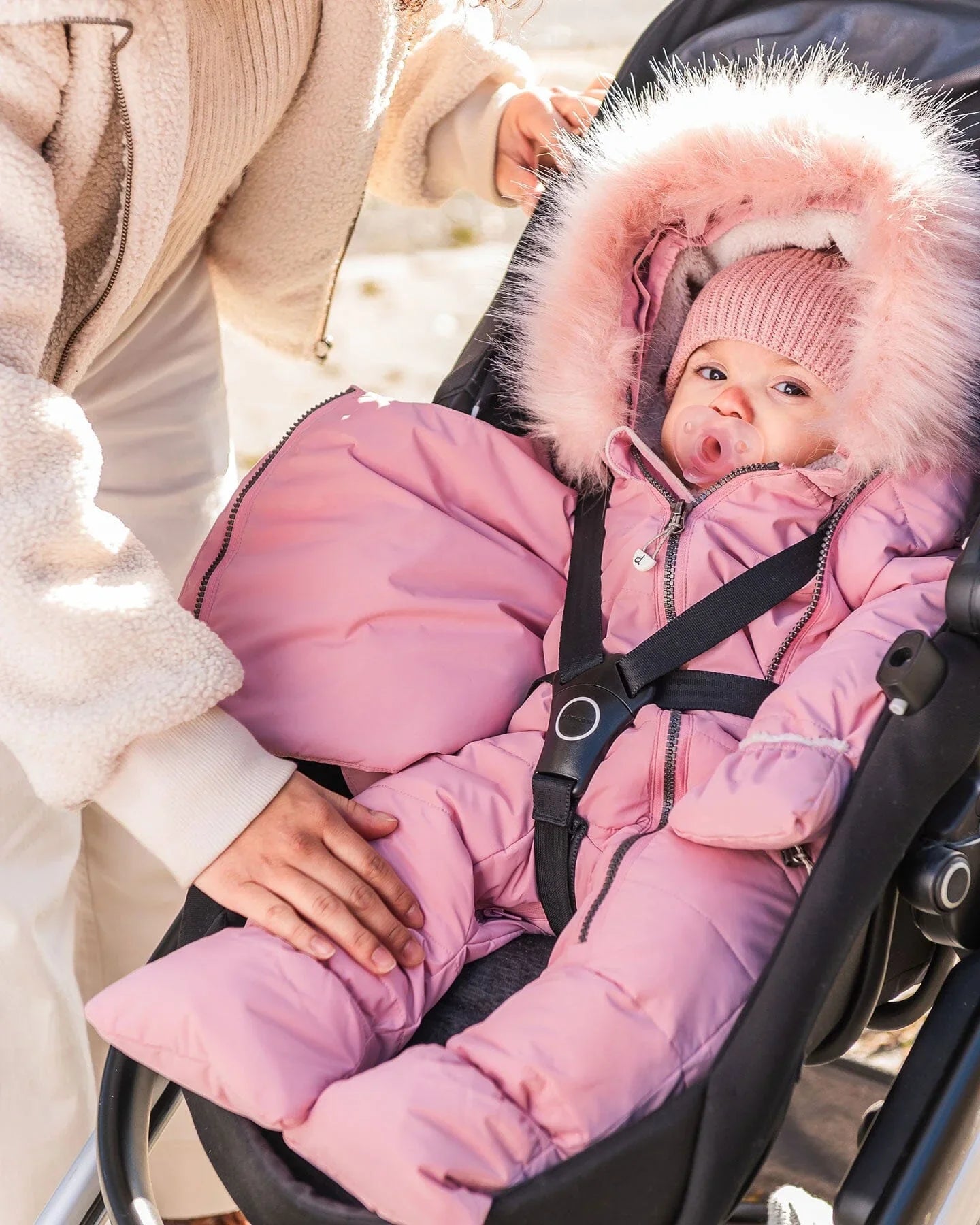 Infant in pink faux fur hooded snowsuit with pacifier in stroller, cozy winter accessories