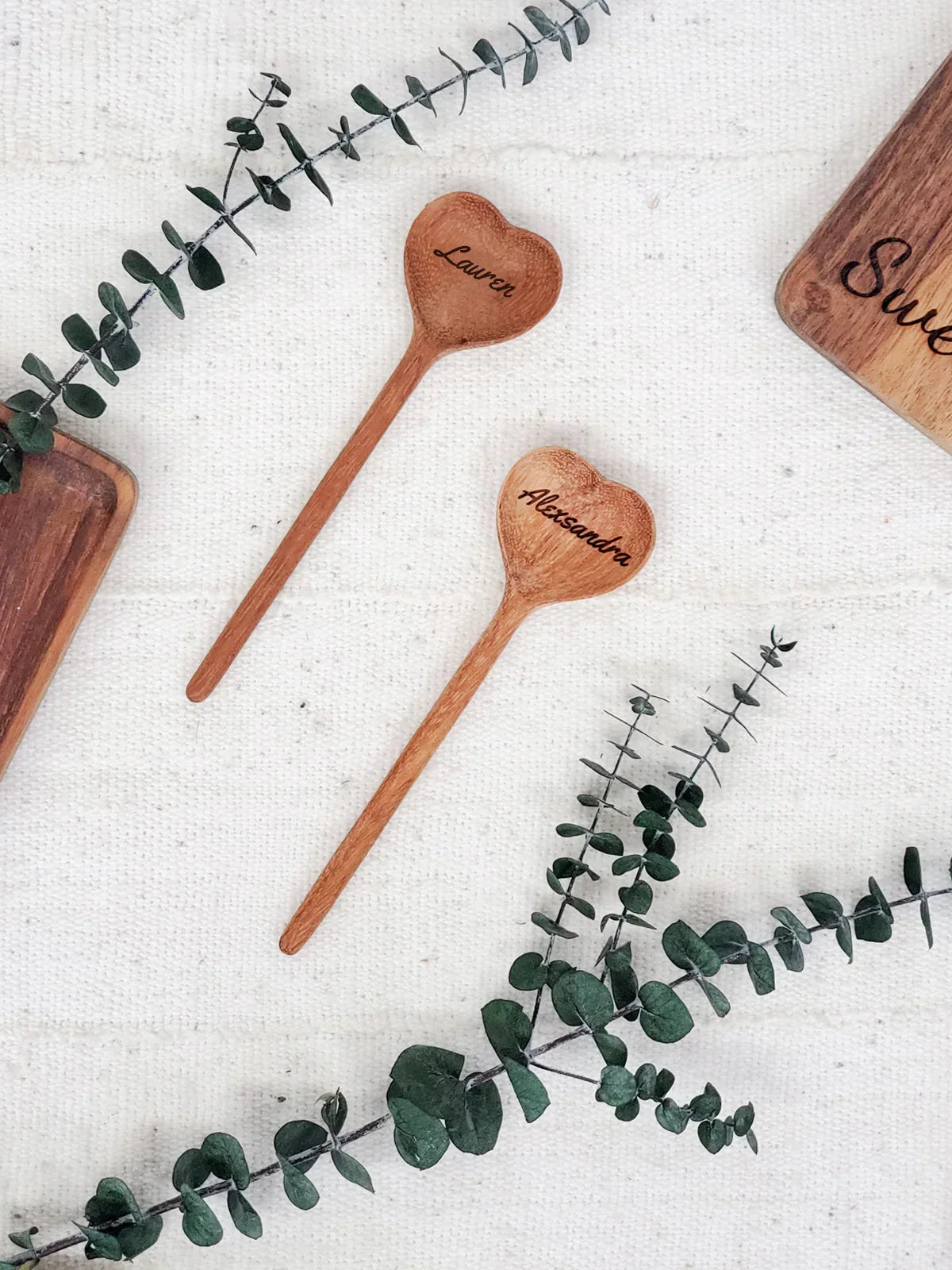 Personalized wooden heart-shaped spoons engraved with names Lauren and Alexandra on fabric with eucalyptus sprigs